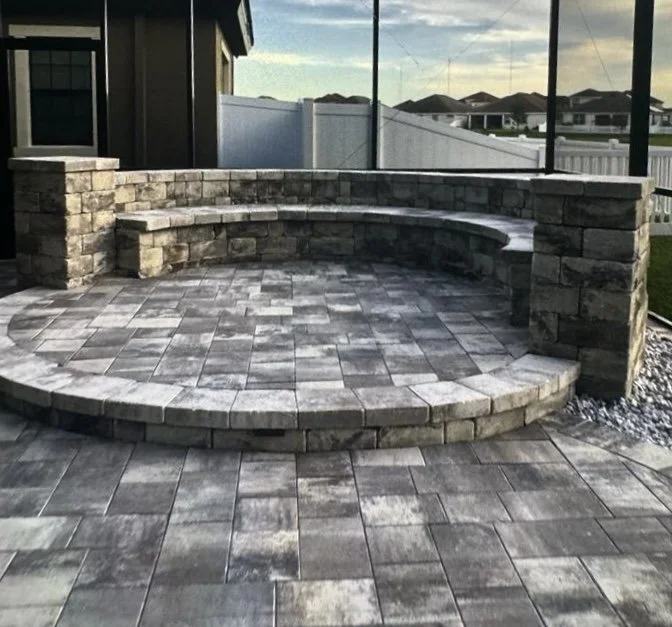 A circular stone patio with built-in curved stone seating, enclosed by a waist-high stone wall, in a backyard near a house, with a white fence and a view of houses and a cloudy sky in the background.