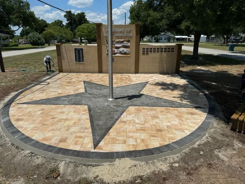 A newly constructed Veterans of Hamilton memorial with a star-shaped design on the ground, a flagpole, and a sign detailing veterans' history, situated in a park with trees and a street in the background.