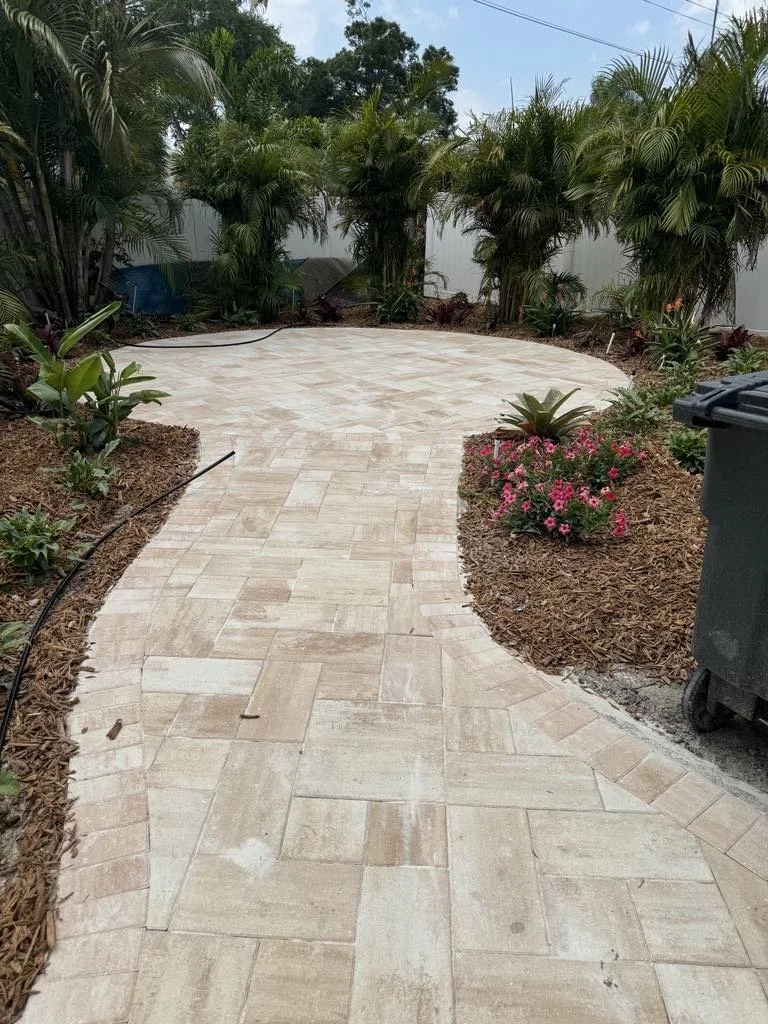 A winding patio pathway made of beige flagstone in a landscaped backyard with tropical plants, pink flowers, a black trash bin, and a white fence in the background.