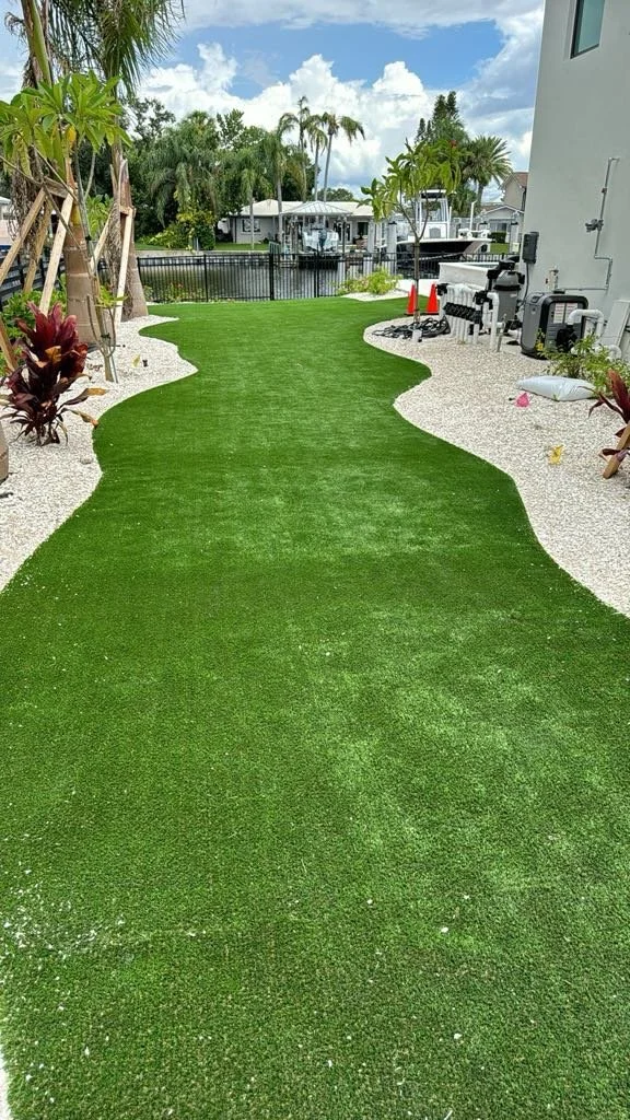 Artificial green grass pathway in backyard with white gravel border and tropical plants on each side, leading towards a canal with boats.