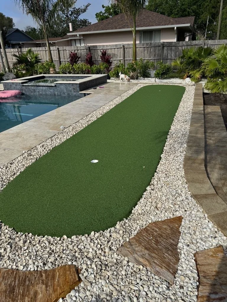 A backyard with a mini golf putting green, surrounded by white rocks and decorative flagstone, next to a swimming pool with a hot tub, trees, and plants in the background.