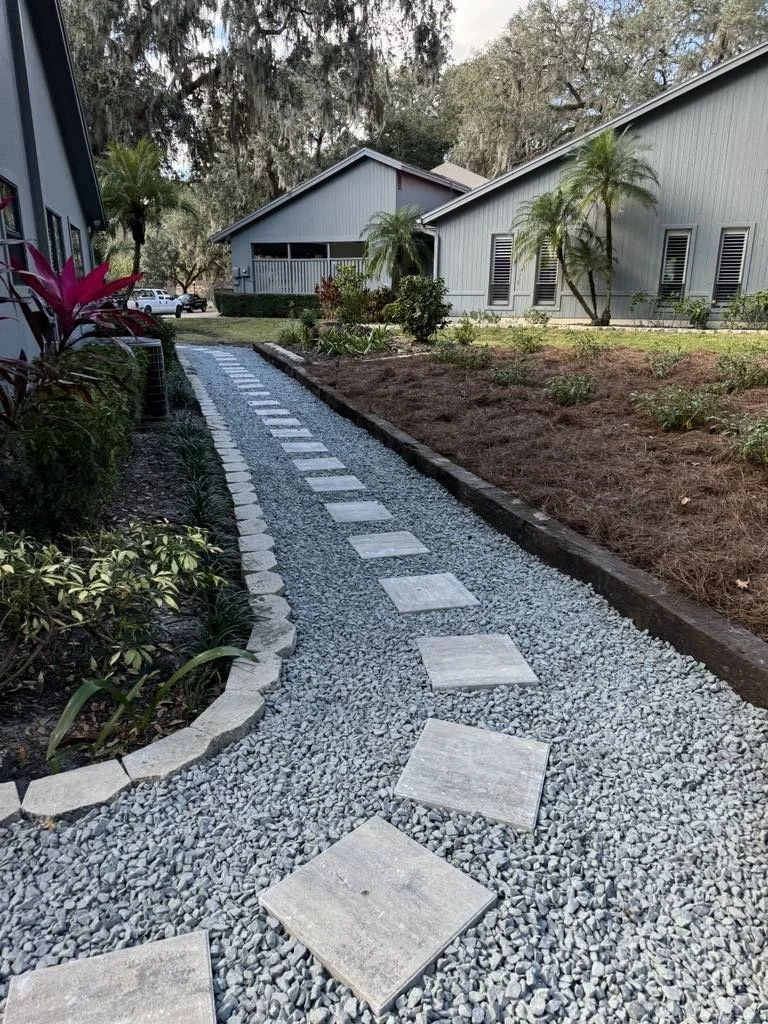 A curved gravel pathway with flat square stepping stones in a garden area between two residential buildings, with plants and trees alongside.