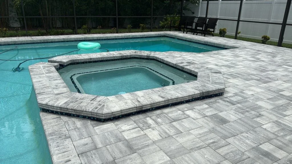 A backyard swimming pool with a hot tub in the foreground, surrounded by a stone patio and enclosed by a screened fence, with outdoor seating and greenery in the background.
