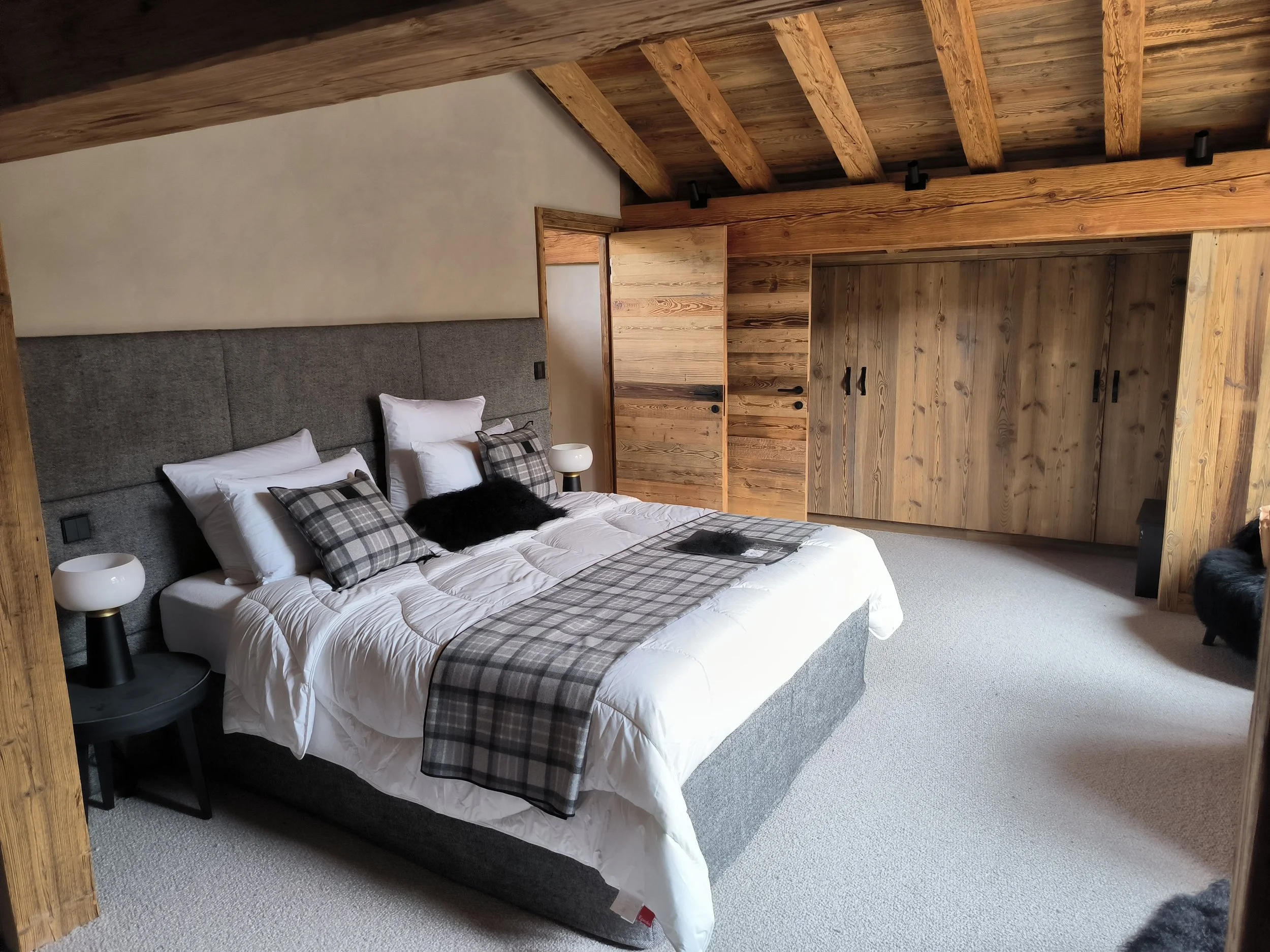 Chambre à coucher avec lit en bois, tête de lit sur mesure en modules rectangulaires tissu gris, draps blancs, oreillers à carreaux, lampe de chevet, murs en bois et plafond en bois avec poutres apparentes.