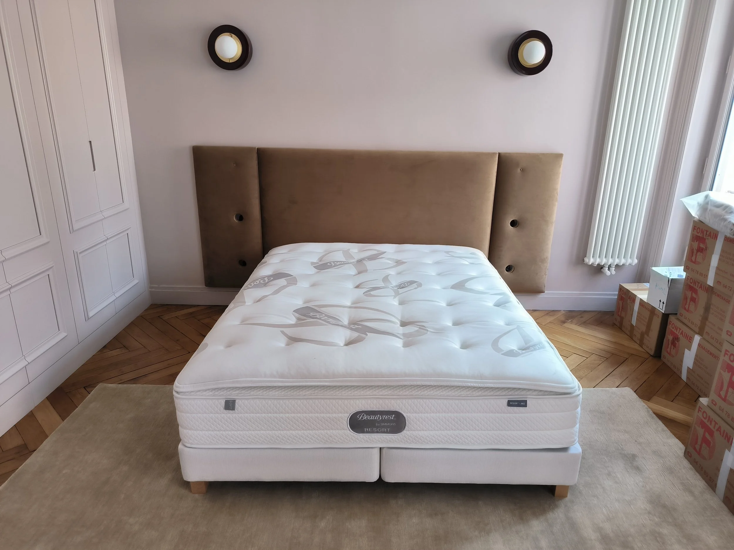 Un lit avec un matelas blanc sans draps, un tête de lit en velours beige et deux appliques murales rondes.