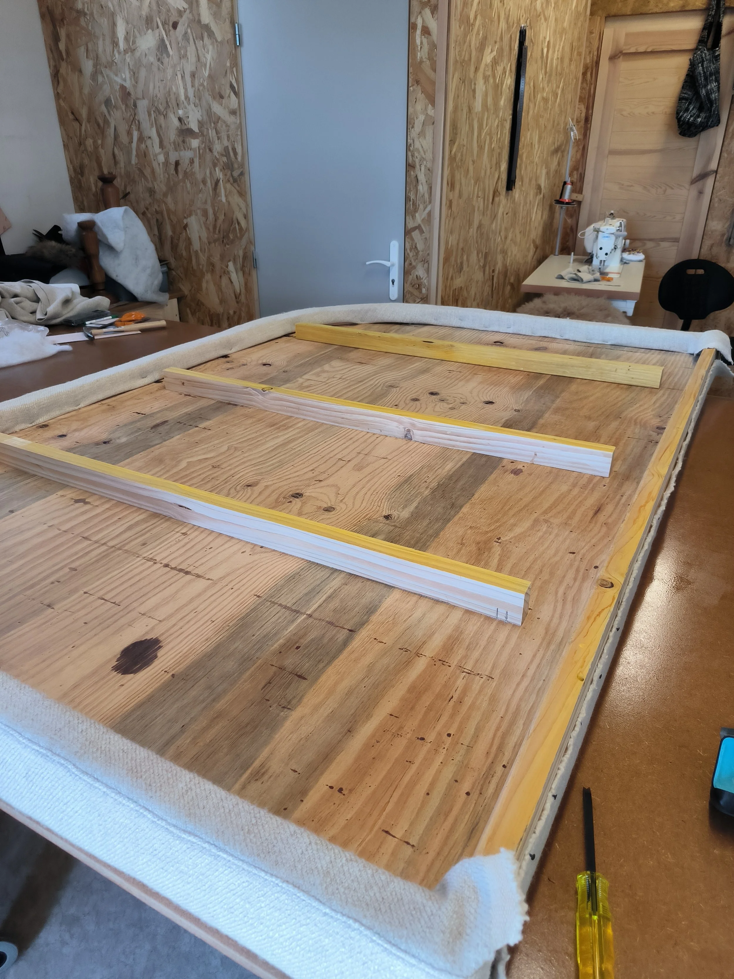 Un atelier de bricolage avec des outils, une planche de bois, et un fond en panneau de bois. On voit une table de travail avec des outils et des matériaux de couture, une machine à coudre, et un fauteuil, dans un environnement de travail ou de atelier.