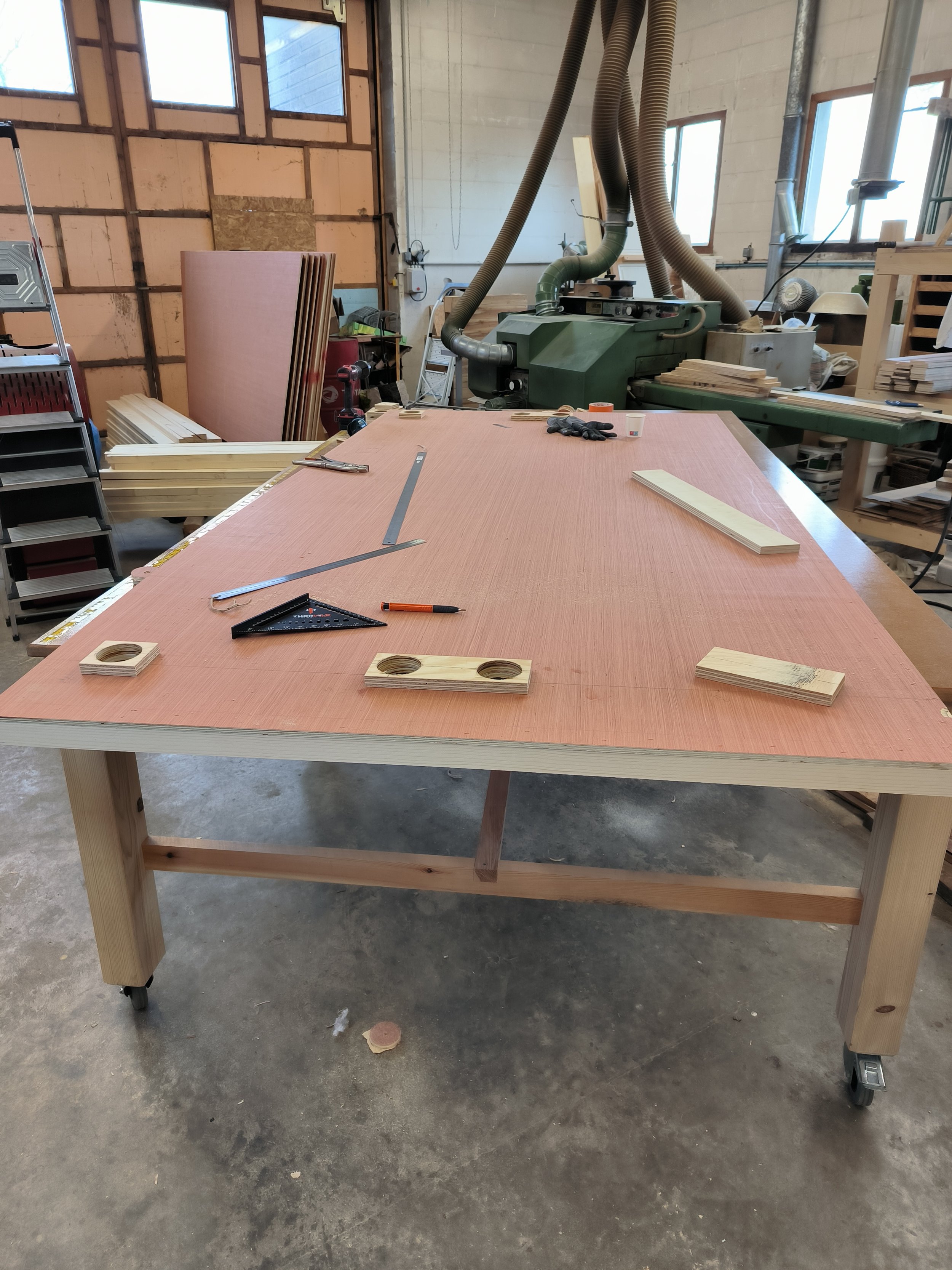 Atelier de menuiserie avec une grande table en bois, outils, pièces de bois, et une machine à découper, dans un espace bien éclairé par plusieurs fenêtres.