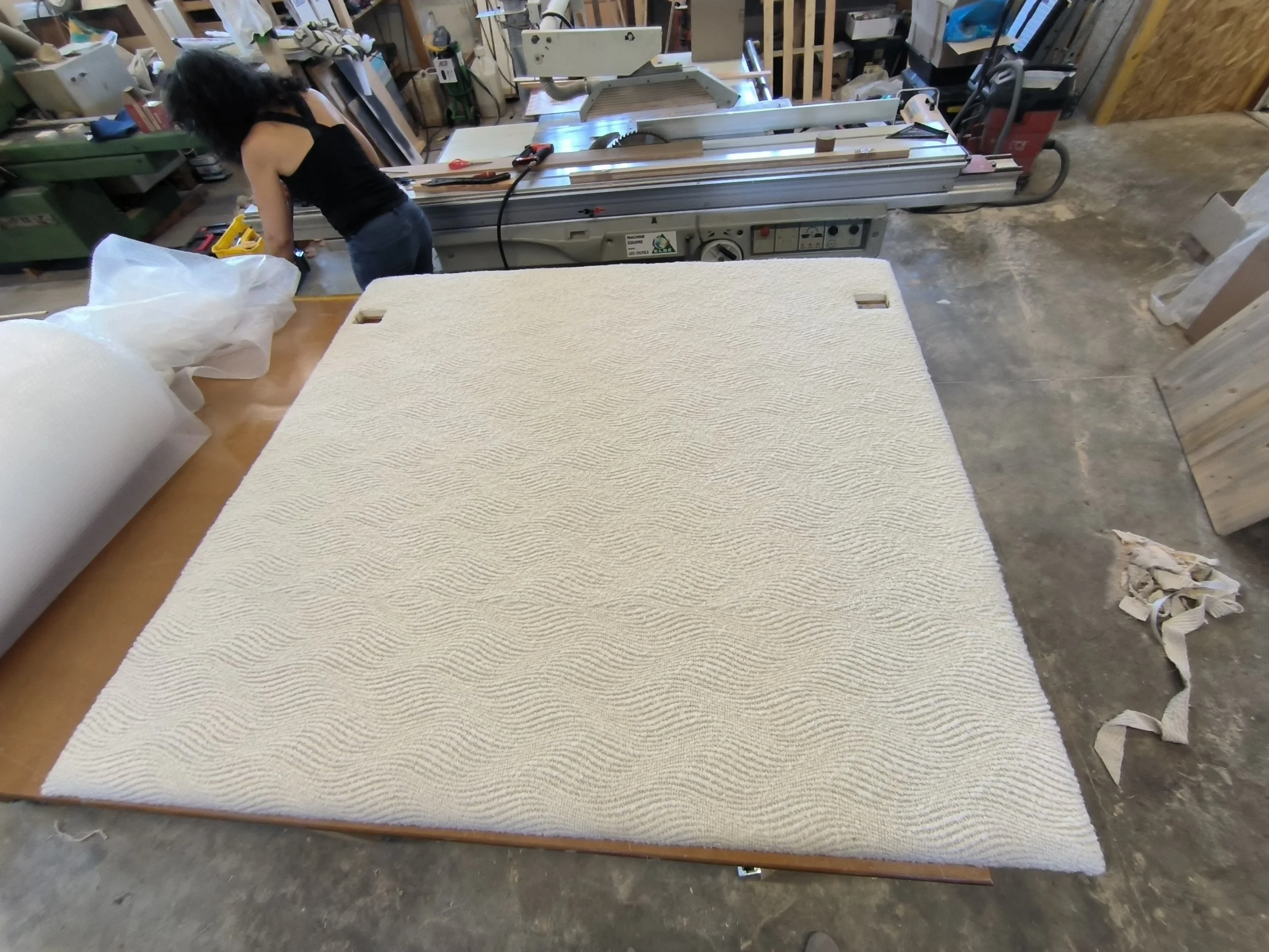 Une personne utilisant une machine à bois dans un atelier, avec une tête de lit recouverte d'un tissu bouclette blanc en premier plan.