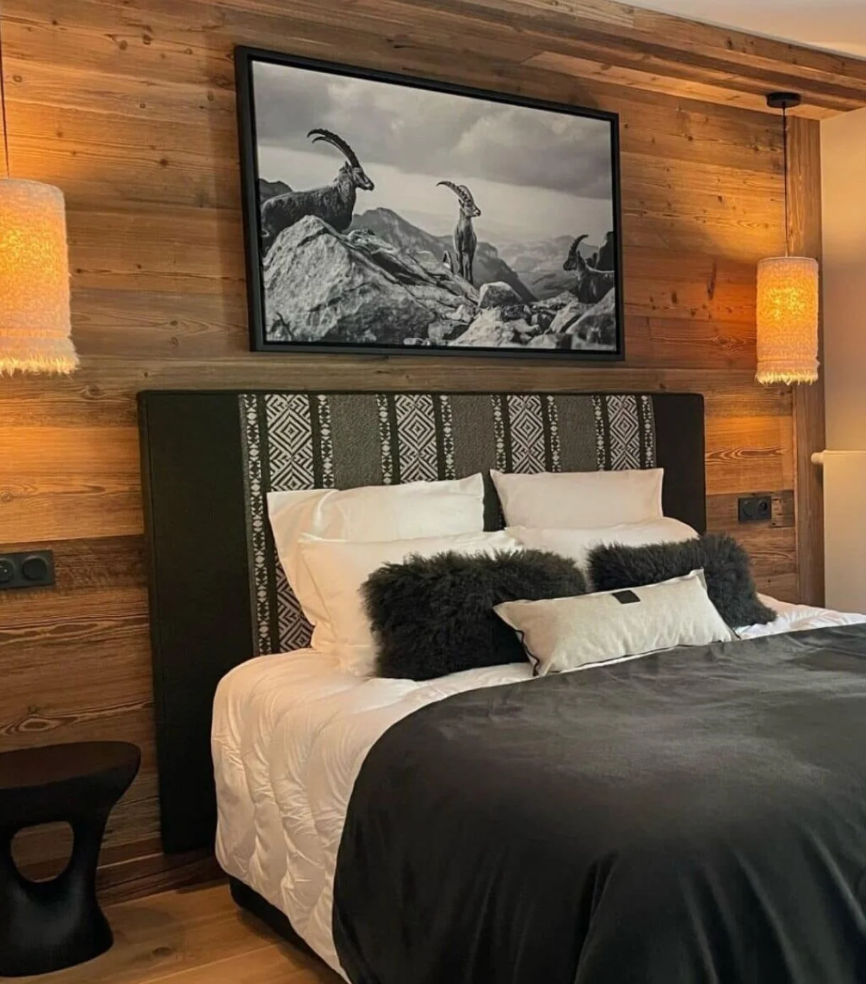 Chambre avec lit double et une tête de lit sur mesure en tissu tribal, intérieur en bois, décorée avec des coussins noirs en fourrure et un tableau noir et blanc de chèvres sur le mur au-dessus du lit, ambiance chaleureuse et moderne.