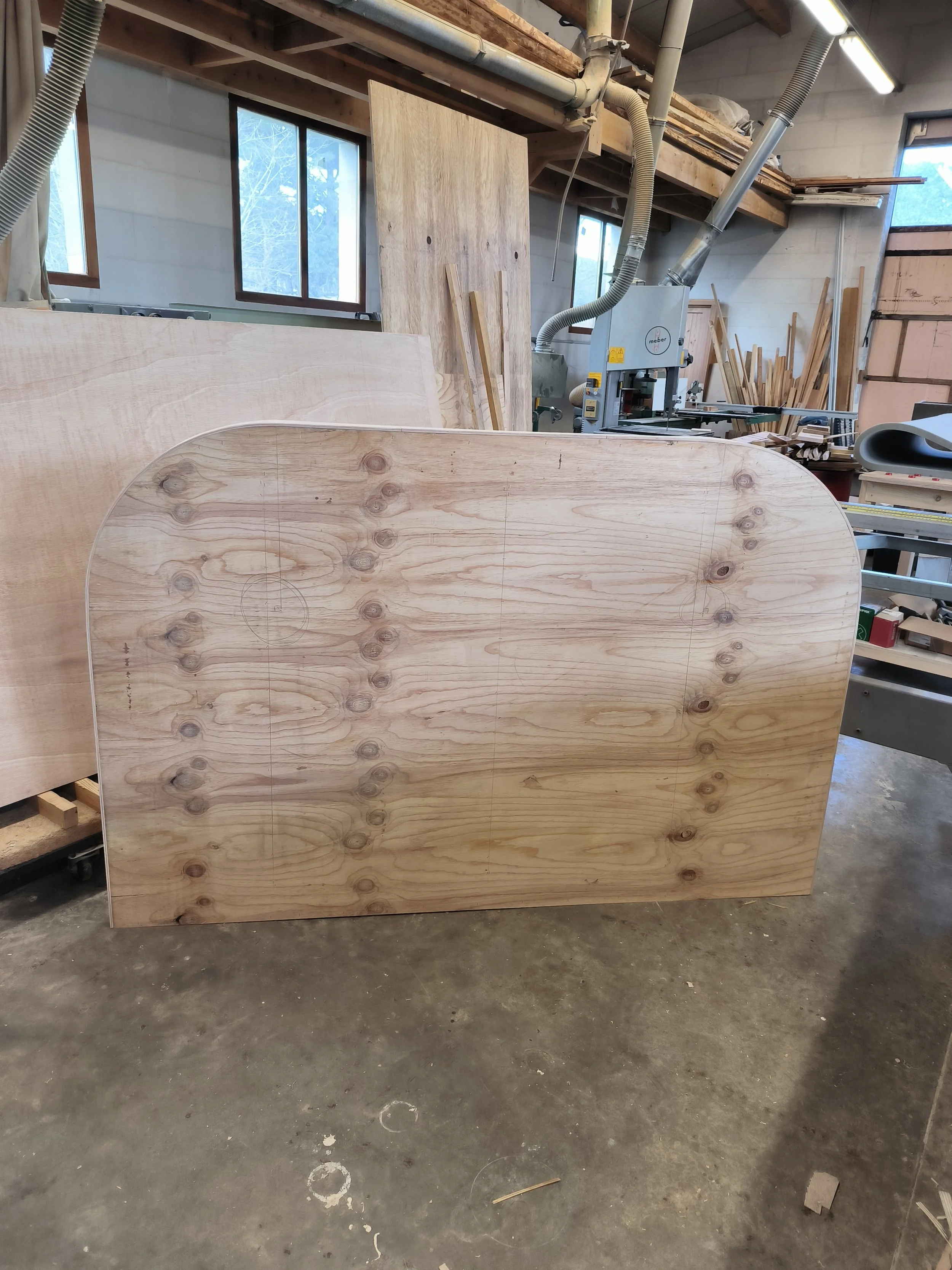 Fabrique d'ossature de tête de lit en bois dans un atelier avec des panneaux en bois, une machine à scier et des fenêtres laissant entrer la lumière naturelle.