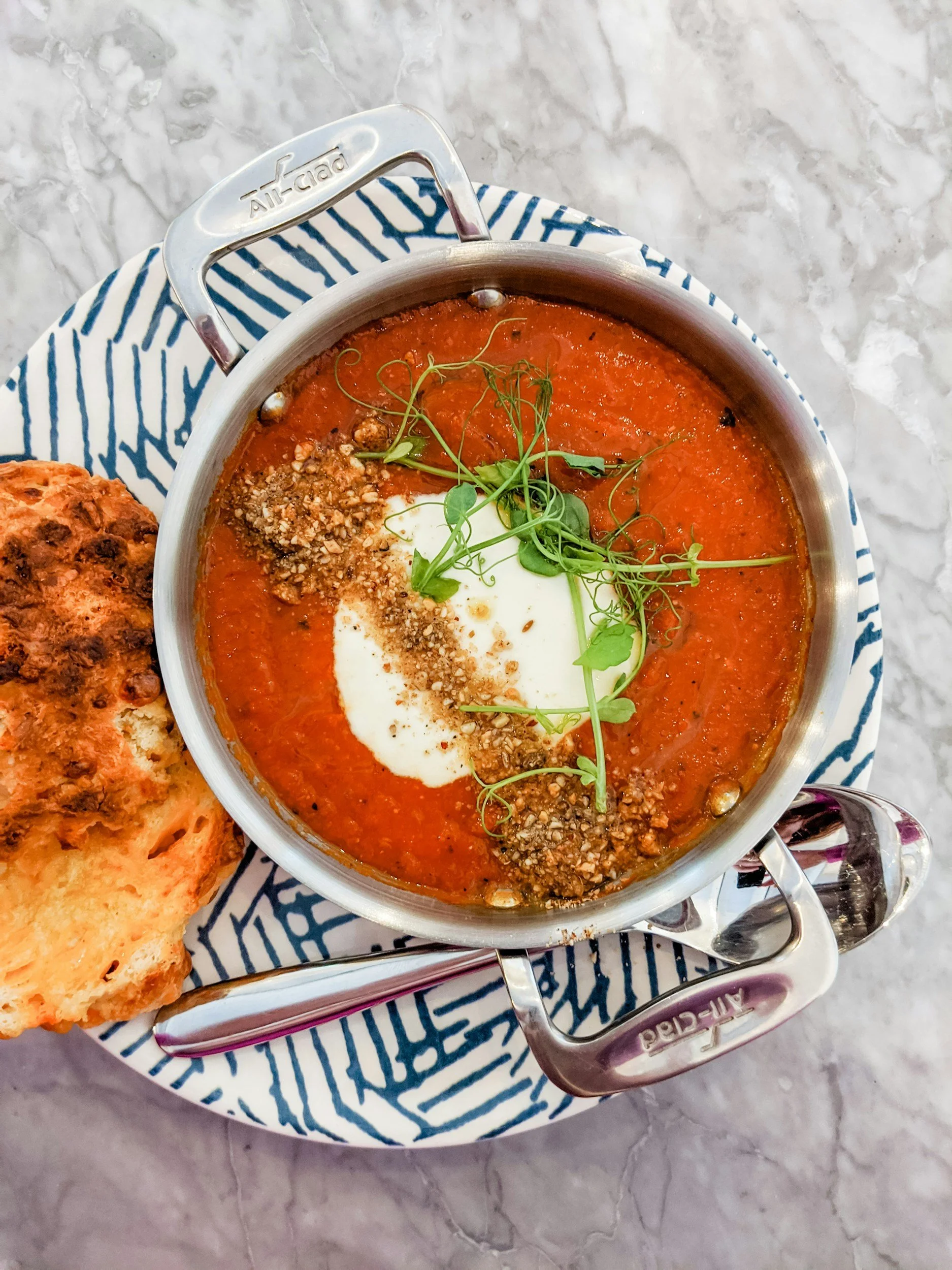 A bowl of tomato soup topped with microgreens, a dollop of sour cream, and chopped nuts, served on a blue and white patterned plate with a biscuit on the side. The setting is on a marble table.