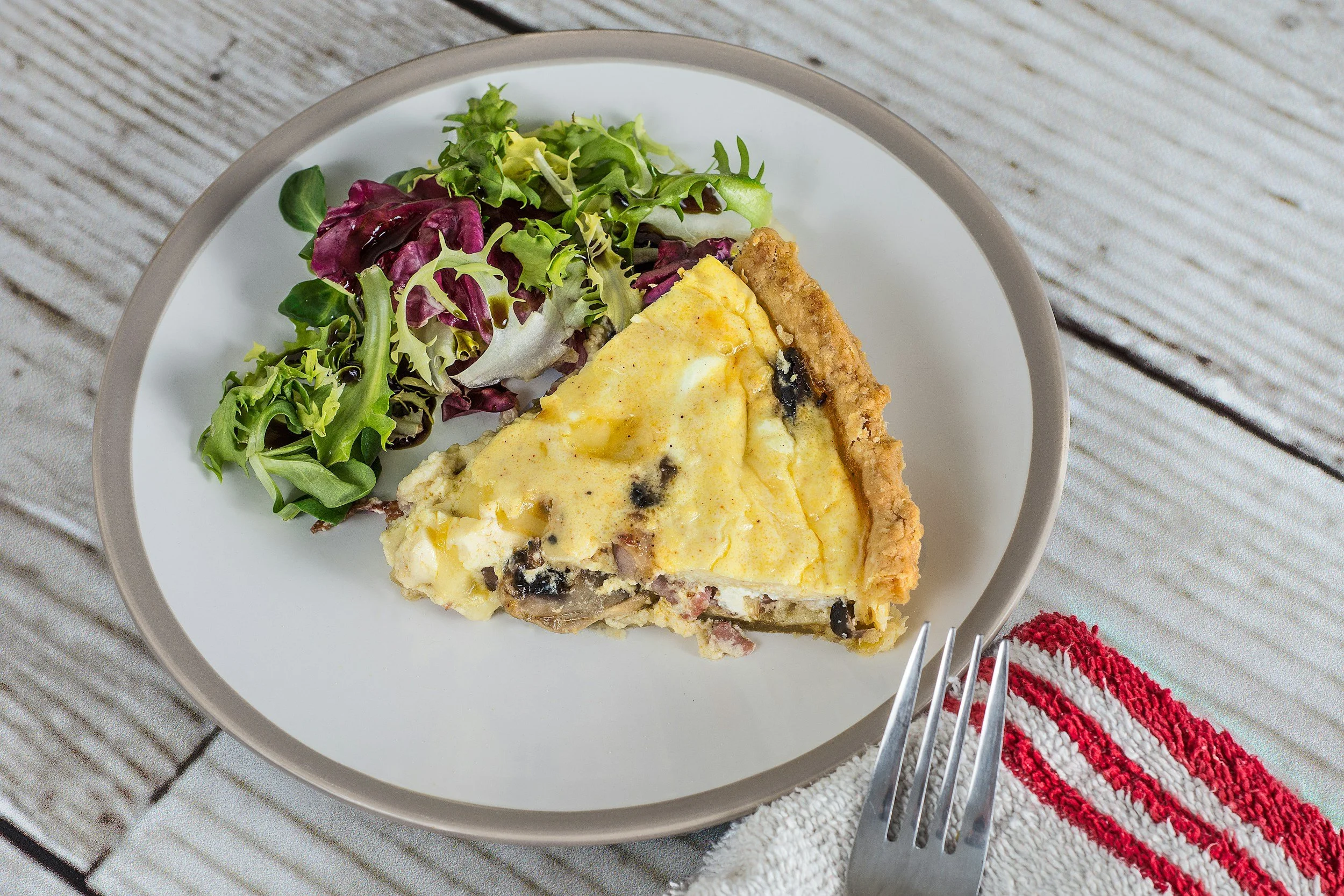 A slice of cherry pie with a buttery crust and lattice topping on a white plate, accompanied by a green salad with mixed greens, on a gray wooden surface with a red-and-white striped cloth and fork.