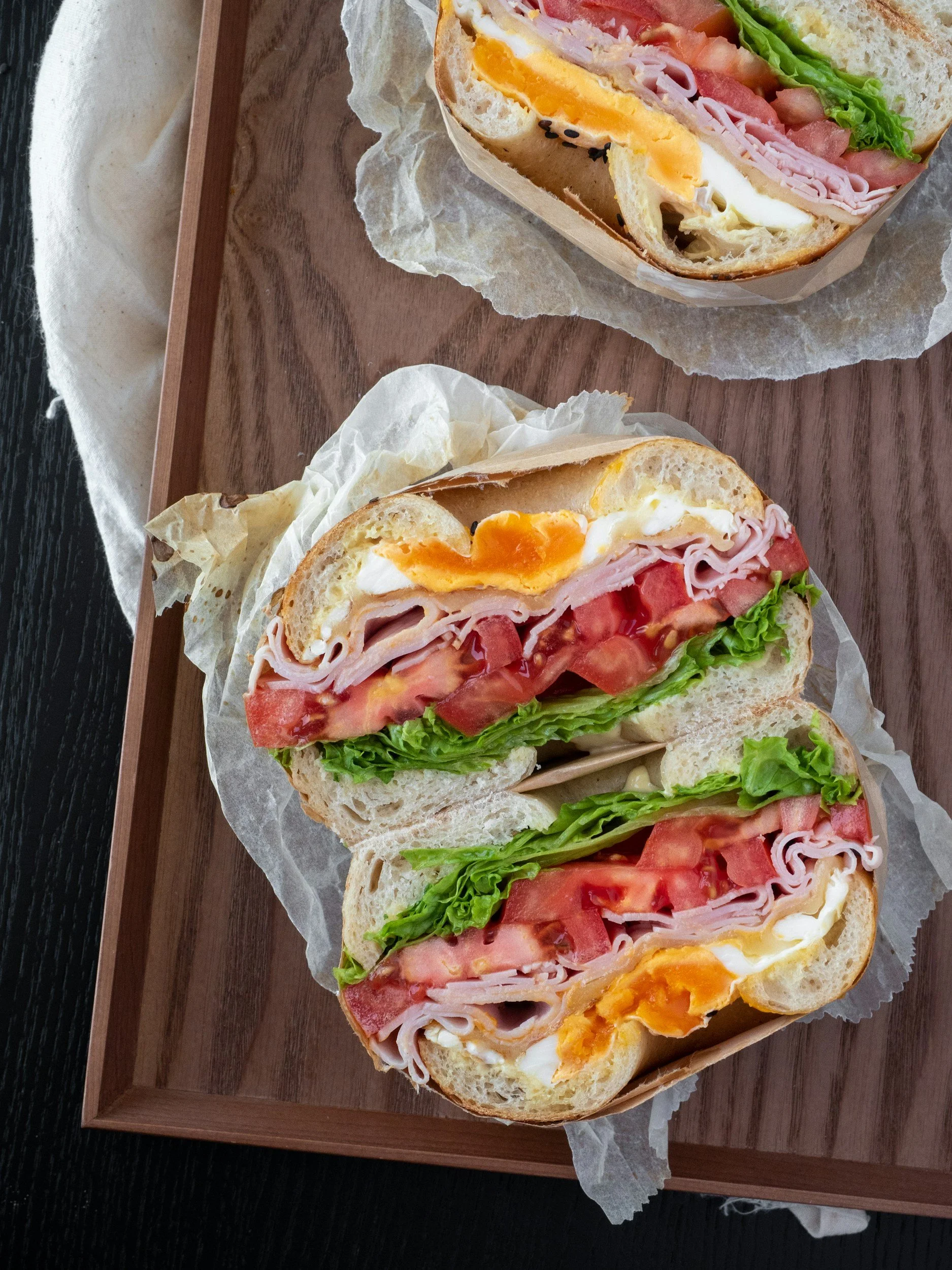 A wooden tray holding three breakfast sandwiches on paper, with ingredients including eggs, ham, bacon, tomato, romaine lettuce, and bread, on a dark surface.