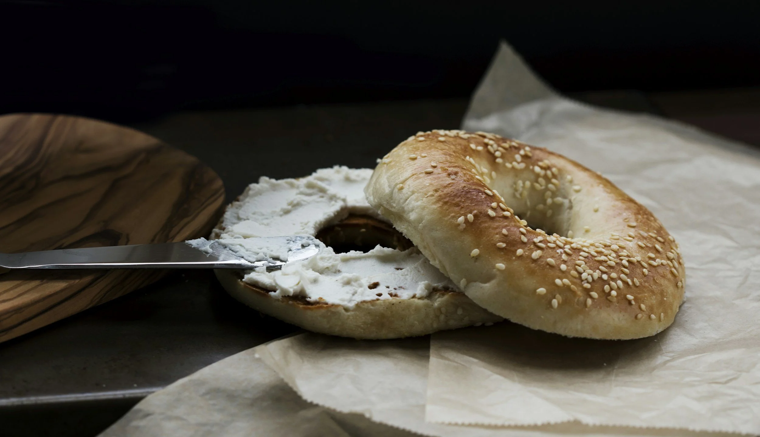 A bagel with sesame seeds on top, partially sliced open with cream cheese spread inside, on a piece of parchment paper, with a butter knife nearby and a wooden cutting board in the background.