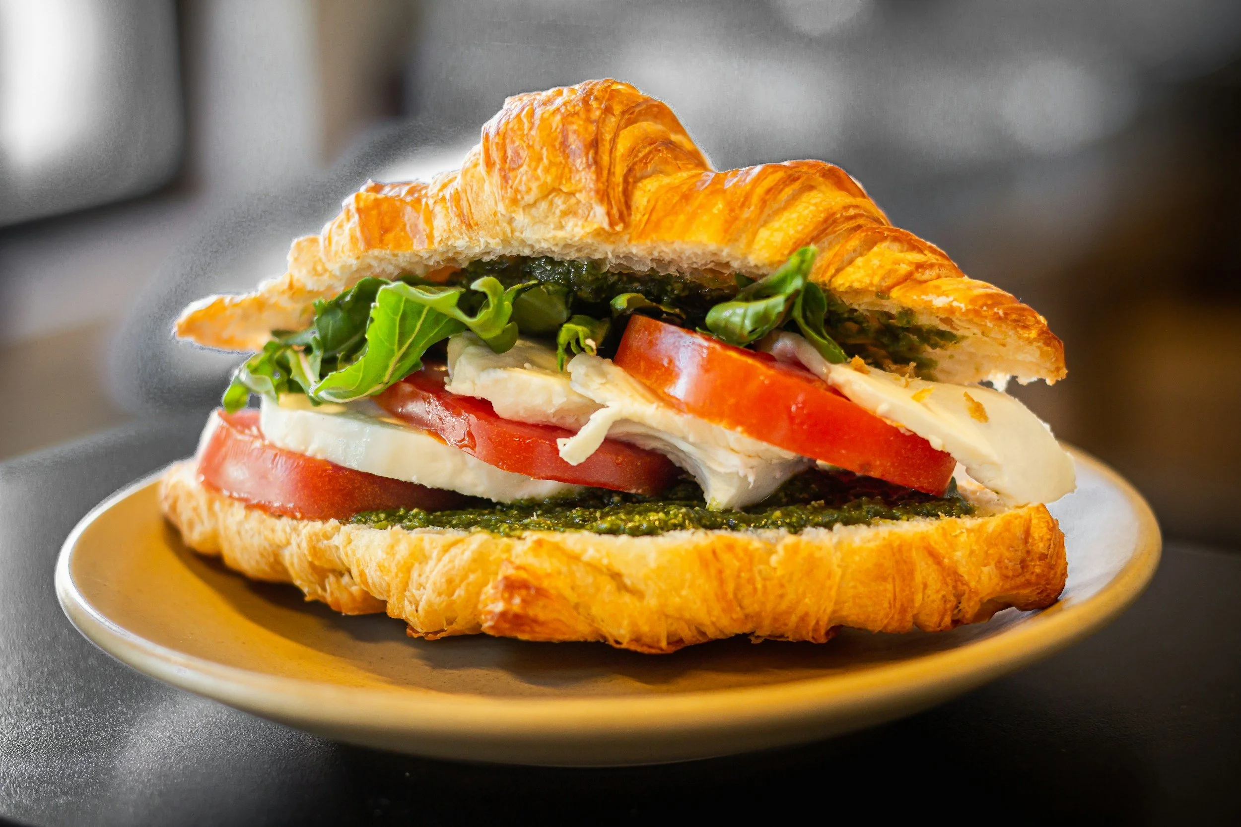 A sandwich with croissant bread, lettuce, tomato, cheese, and a green spread, served on a yellow plate.