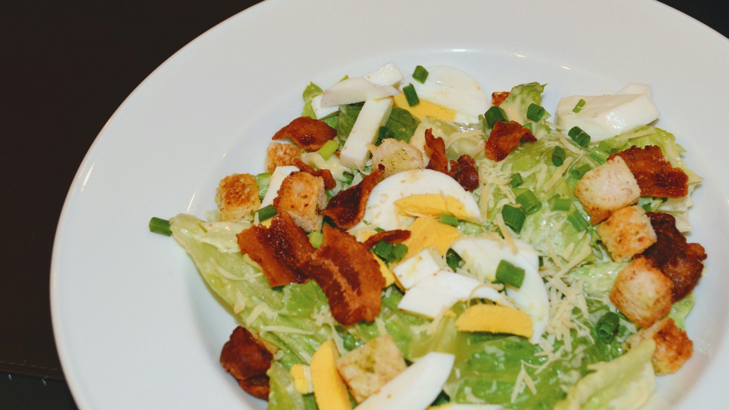 Lettuce salad with sliced hard-boiled eggs, croutons, chopped green onions, shredded cheese, and bacon bits on a white plate.