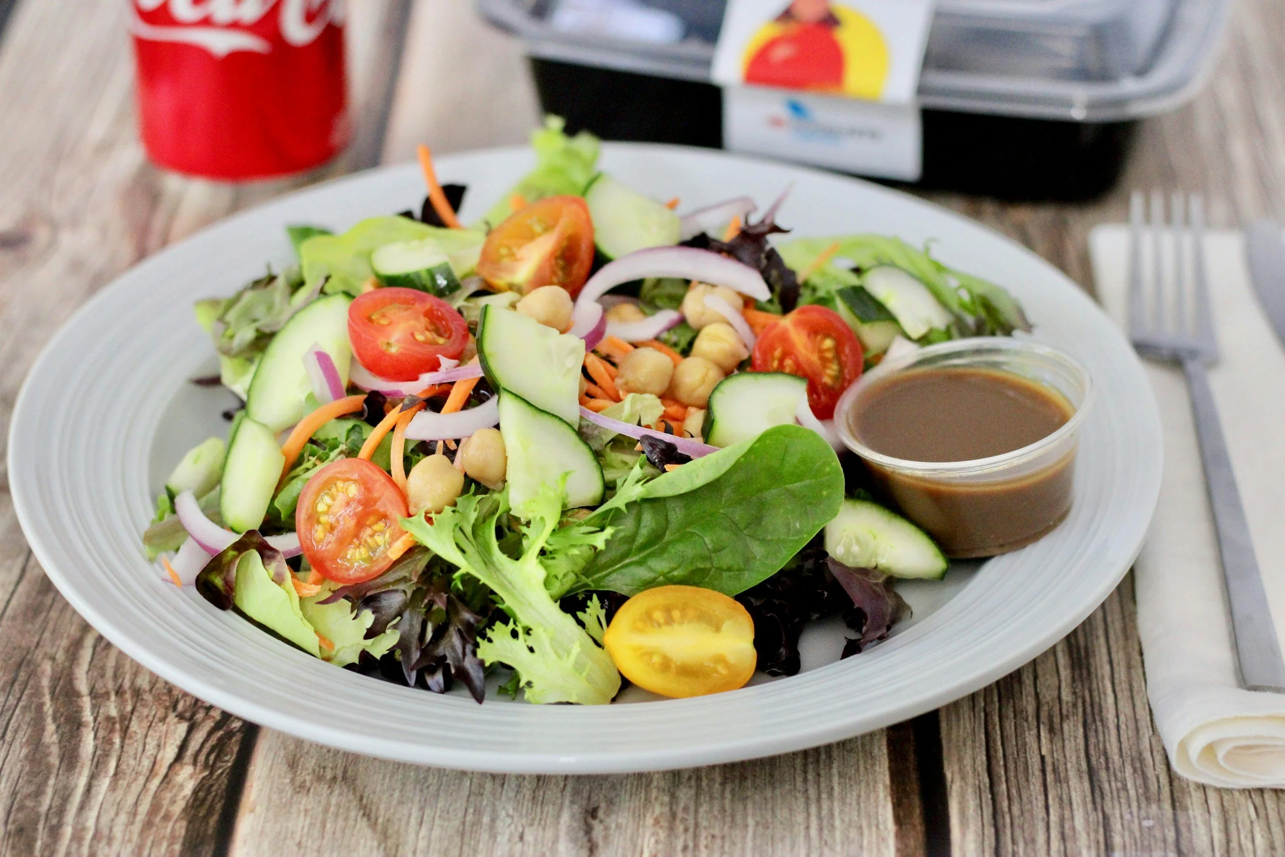 A fresh mixed vegetable salad on a white plate with cherry tomatoes, cucumber slices, shredded carrots, chickpeas, and leafy greens paired with a small container of salad dressing, set on a wooden table.