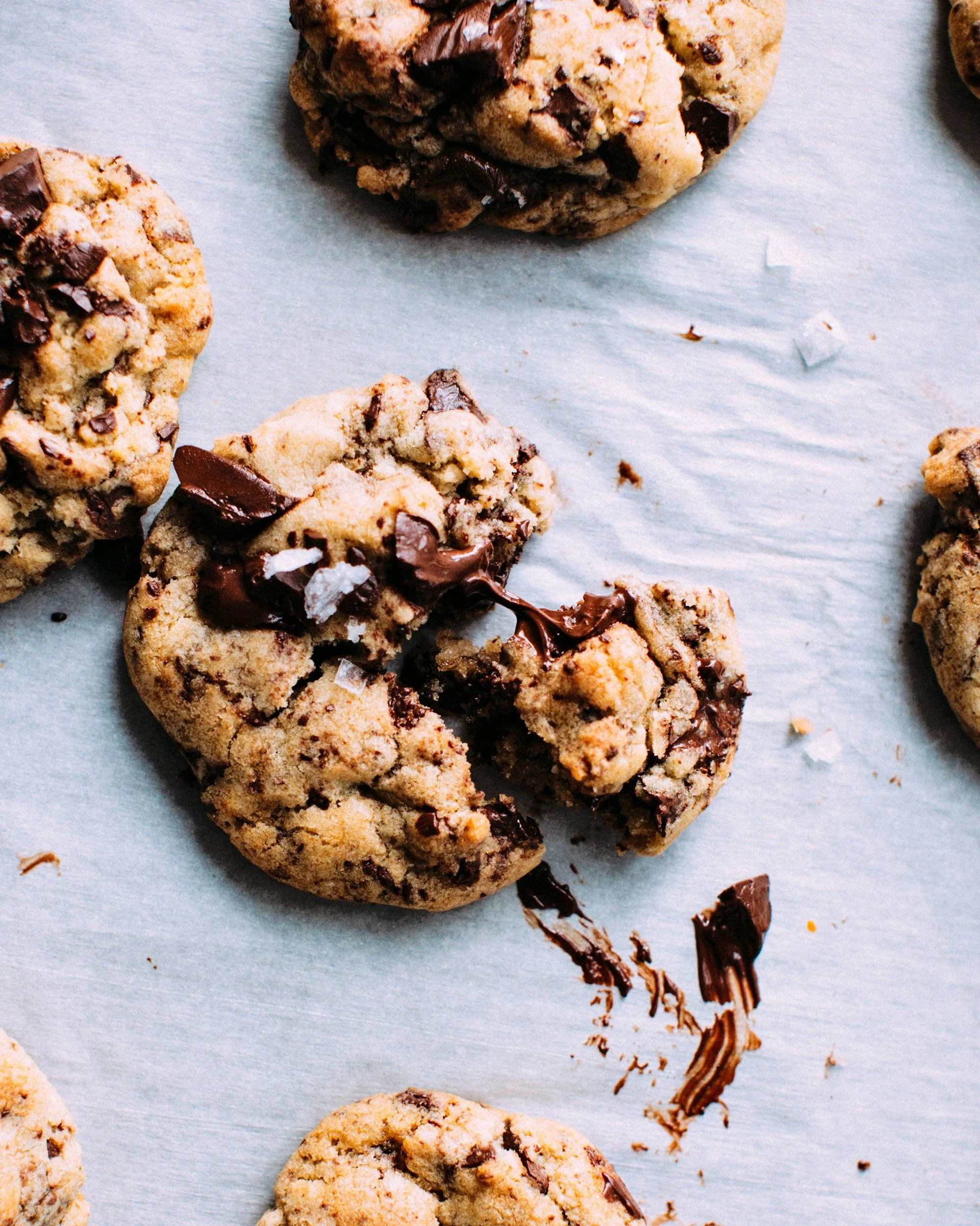 Close-up of several chocolate chip cookies with melted chocolate and some broken pieces on a light-colored surface.