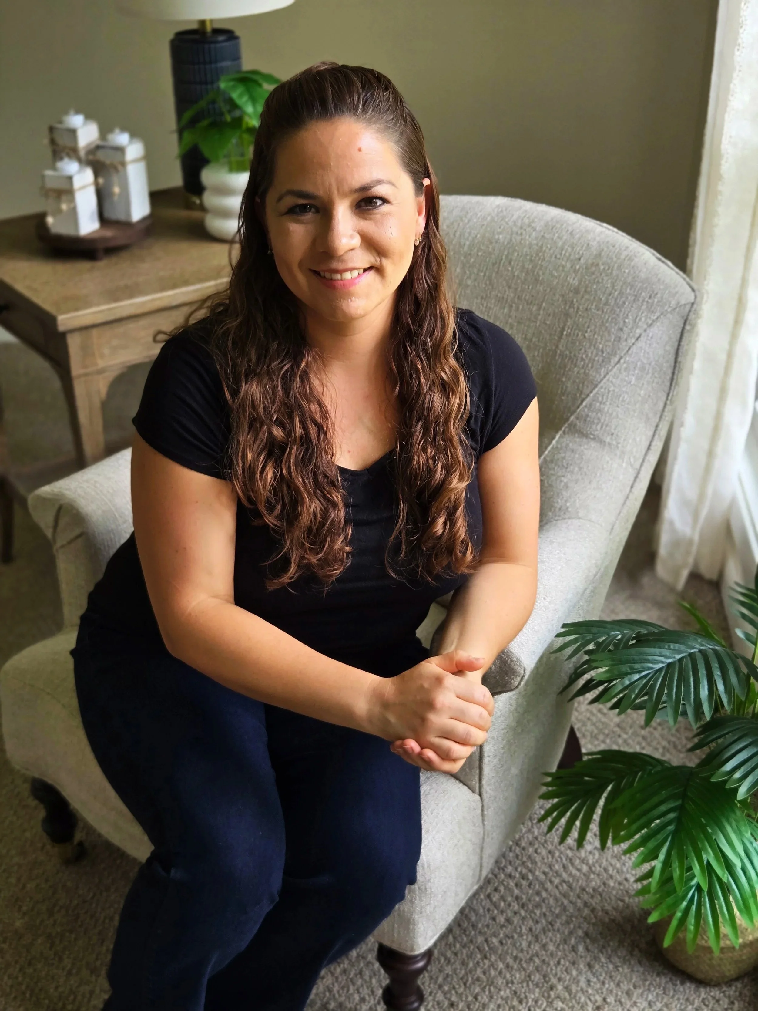 A woman with long curly hair, wearing a black shirt, sitting on a beige armchair in a living room, smiling at the camera with her hands clasped in her lap. A wooden side table with decorative candles and a green potted plant is behind her. There are additional plants visible on the floor and a beige curtain on the right.