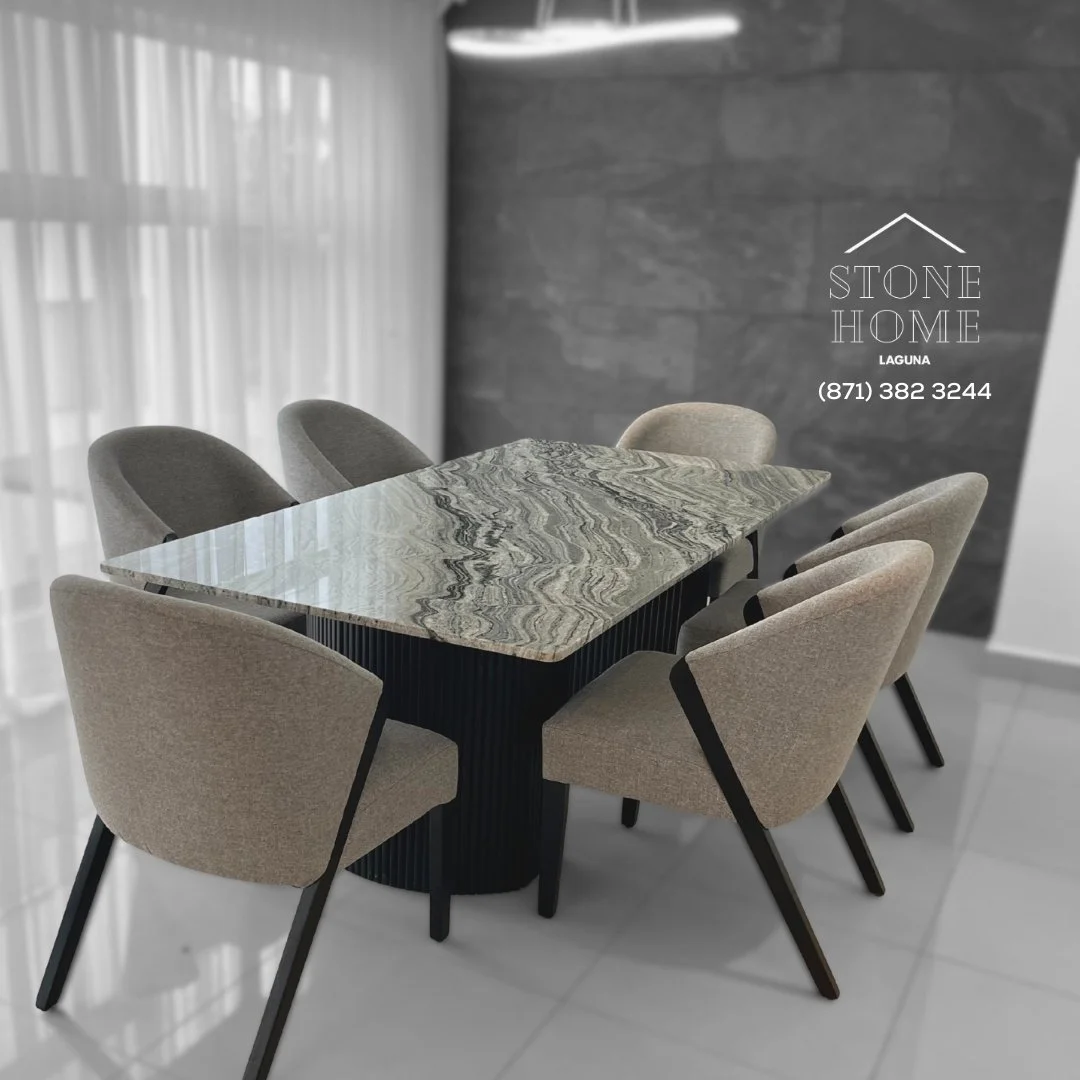 Mesa de comedor con superficie de mármol y seis sillas beige con patas negras en un espacio moderno con cortinas blancas al fondo y pared de piedra gris.