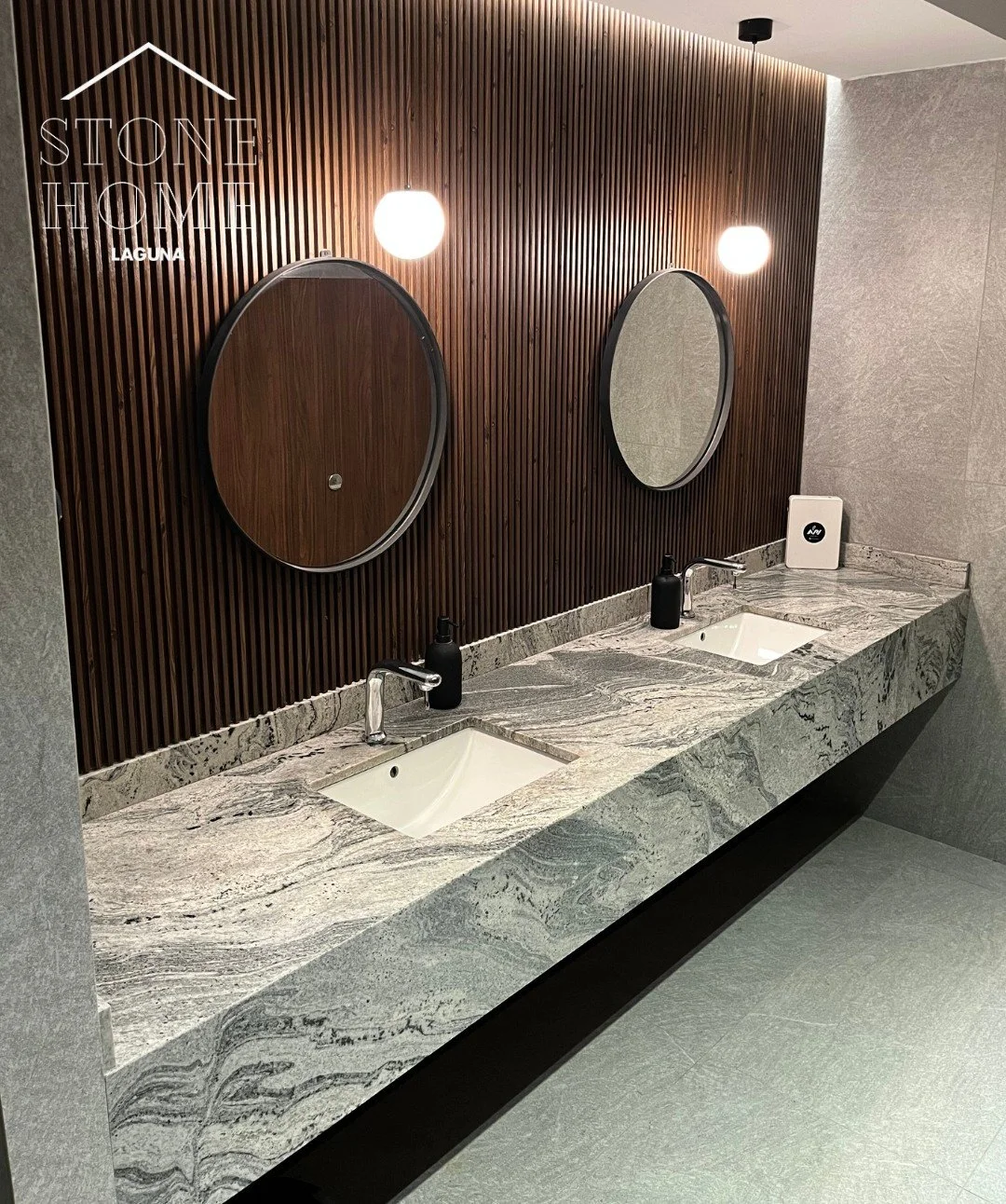 Lavabo de piedra con tres lavabos y espejos redondos en una pared de madera, iluminada con dos lámparas redondas.