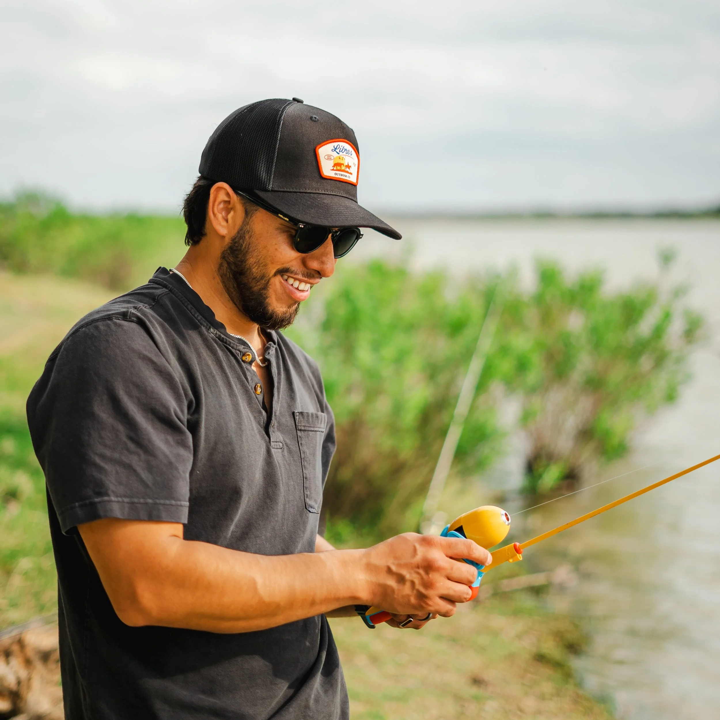 A man wearing a black Libres Outdoors trucker hat with a logo, sunglasses, and a black shirt, smiling while fishing outdoors with a bright, cloudy sky in the background.