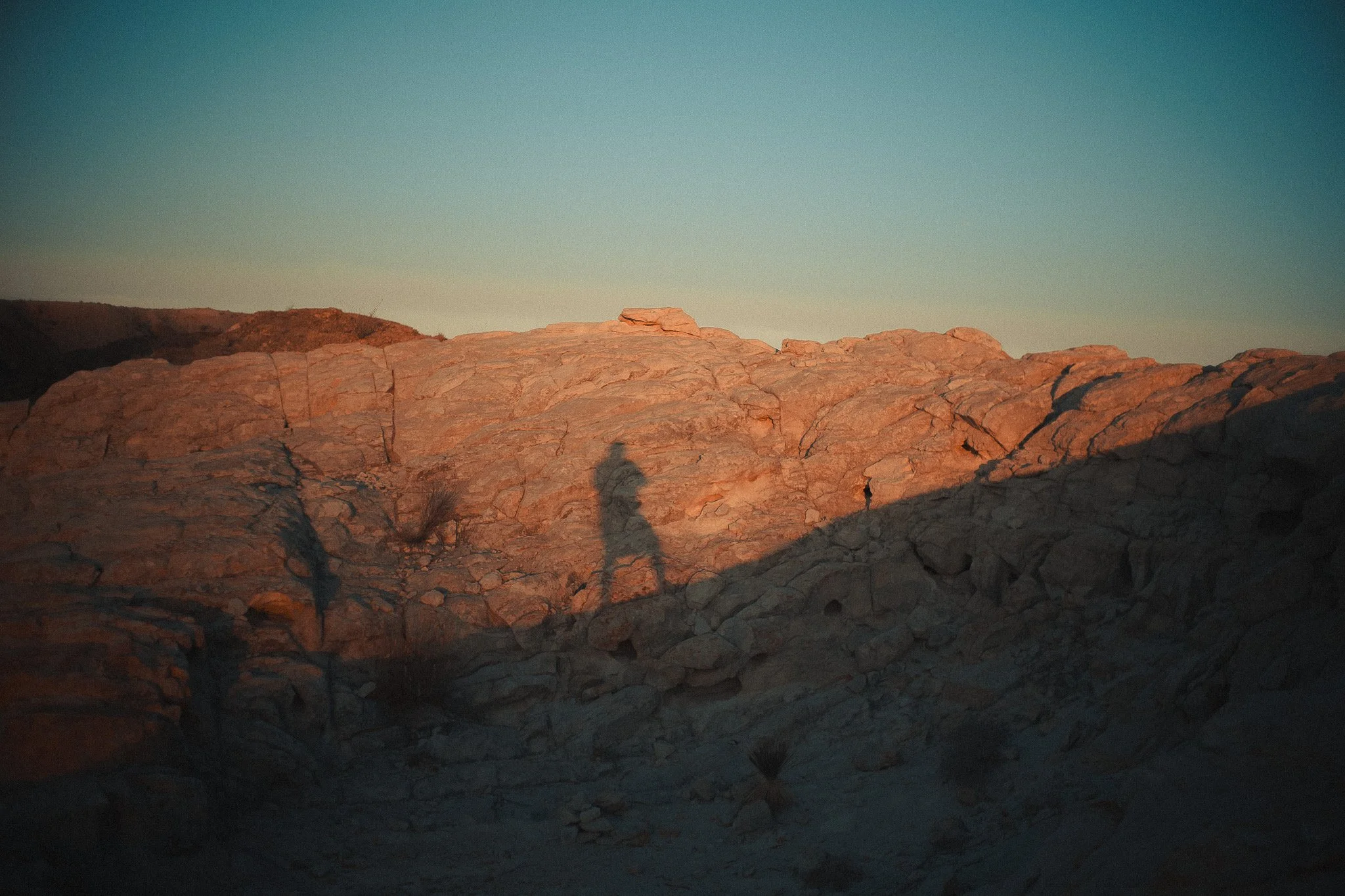 A landscape of rocky desert terrain with low hills in the distance, under a clear sky at sunset or sunrise, with shadows of a person taking the photo visible on the ground.