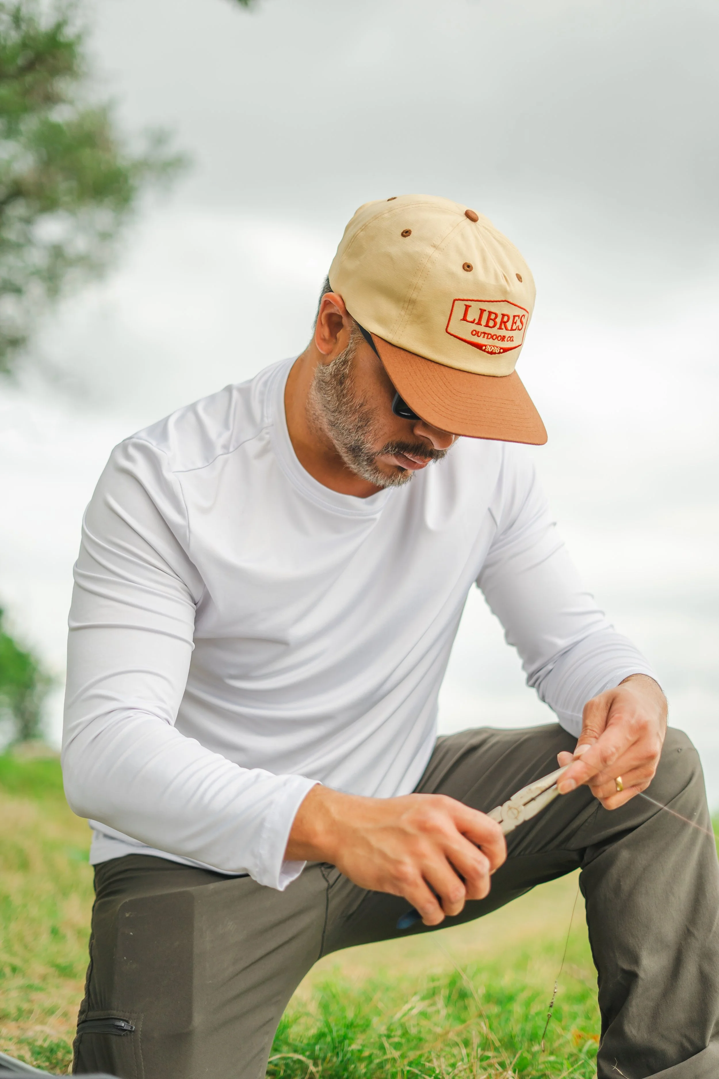 A man in a white long-sleeve shirt and gray outdoor pants crouches outdoors, reading a book under an overcast sky, wearing a tan unstructured hat with red embroidery that says Libres Outdoors.