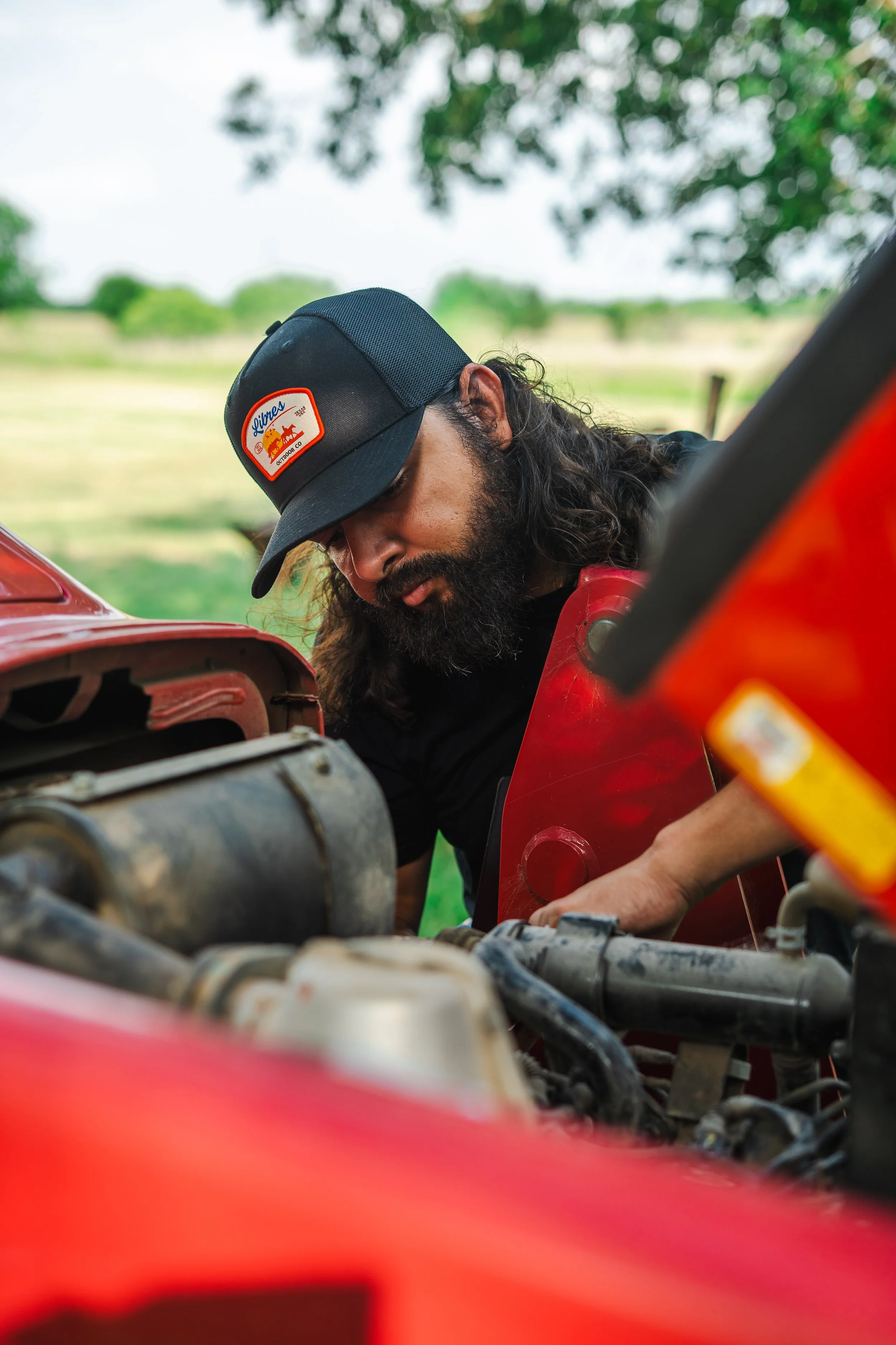 A man with long hair and a beard wearing a black cap with a Libres Outdoors logo, working on the engine of a red vehicle outdoors in a rural setting with trees and open fields in the background.