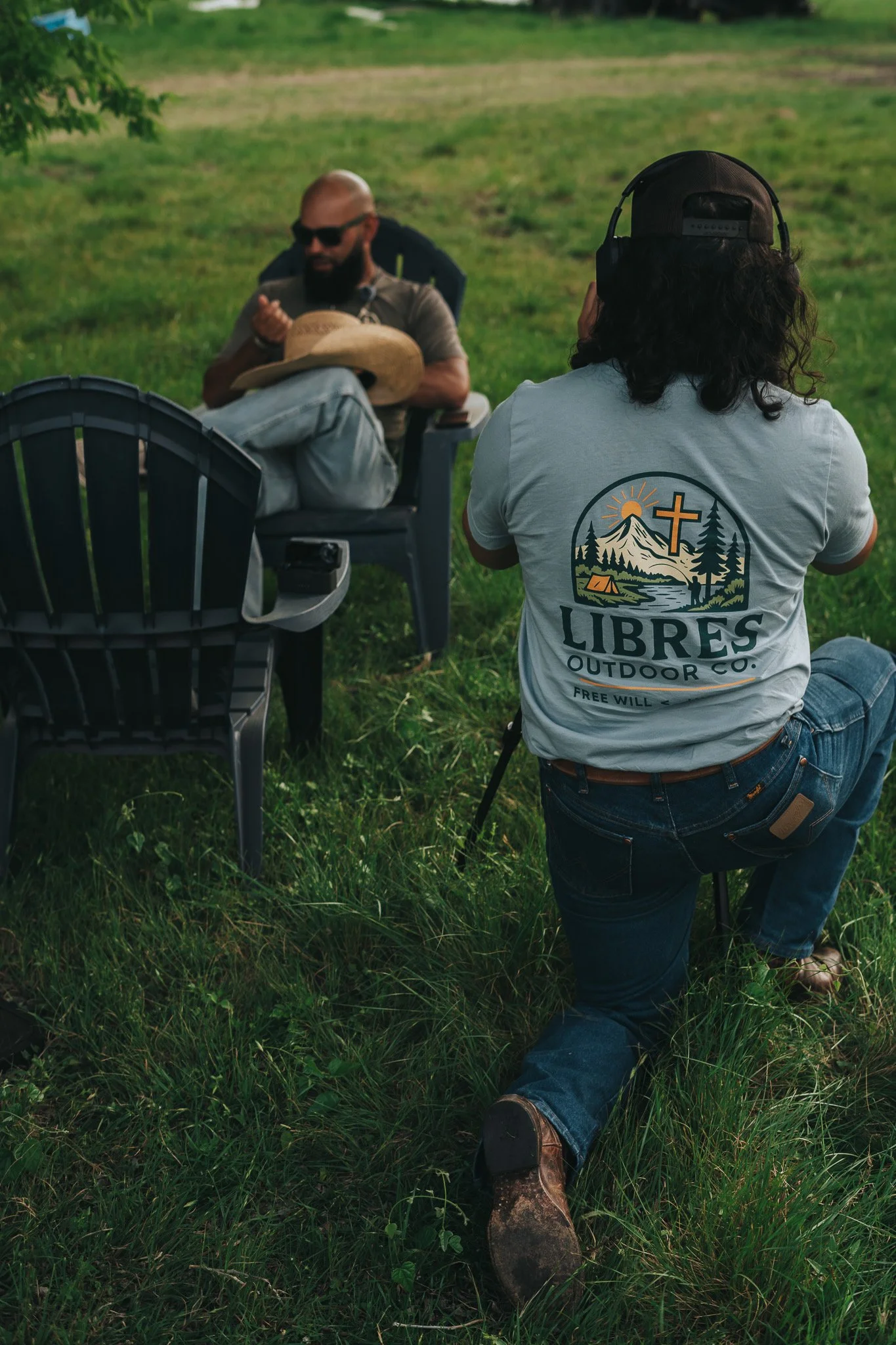 A man is sitting on the grass with his legs stretched out, wearing jeans and brown boots, while a woman kneels on the grass facing him, wearing jeans, a baseball cap, headphones, and a white t-shirt with a nature-themed logo that says 'Libres Outdoor Co.'