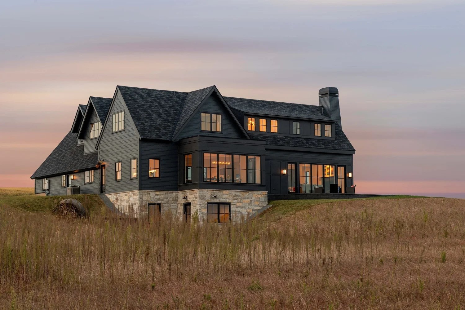 A large, dark-colored house with multiple gabled roofs and many windows sits on a grassy hill at sunset. The house features a stone foundation and bright interior lights are visible through the windows.