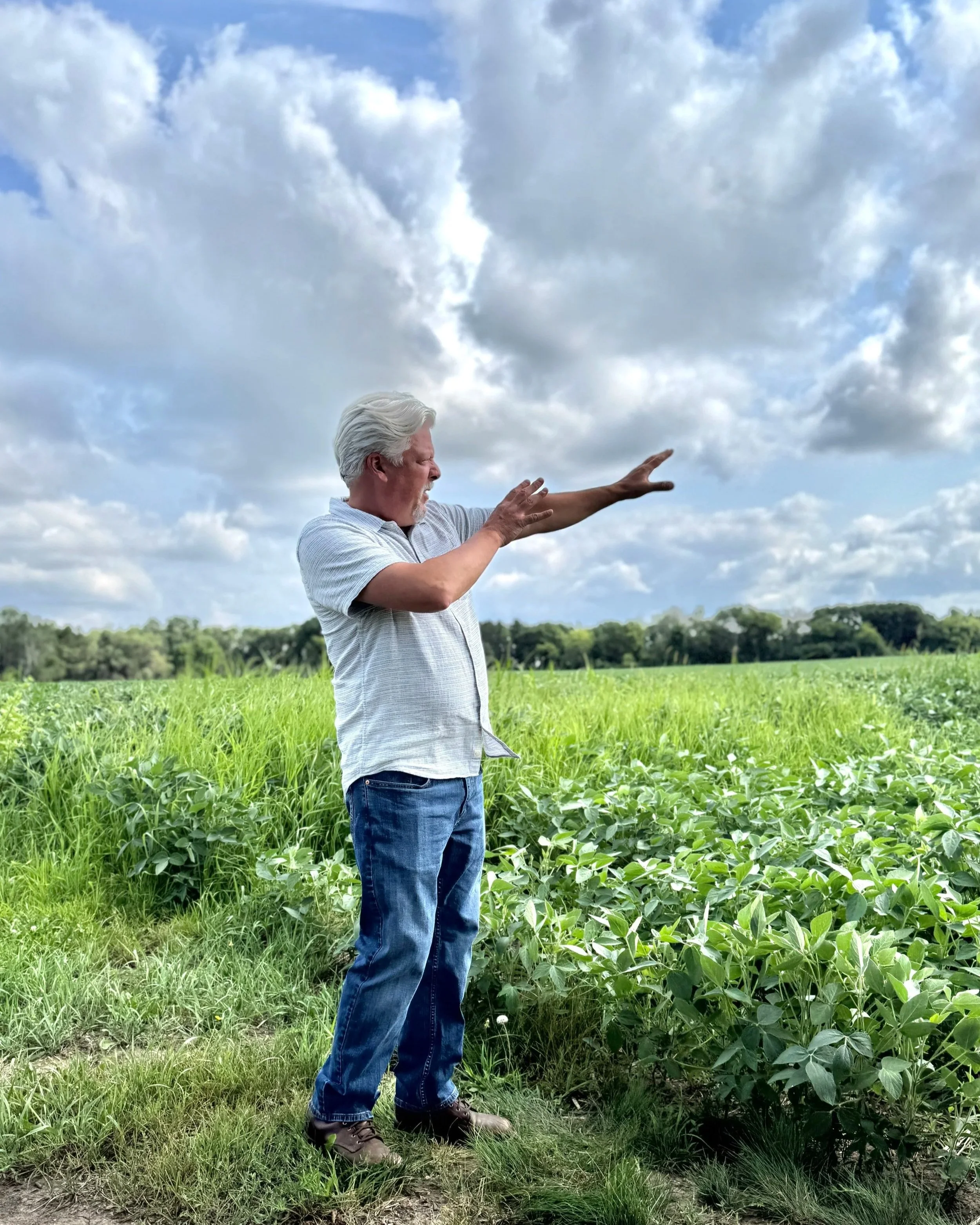 Bob, a man with gray hair and a beard, stands in a field of green crops, wearing a light gray shirt and blue jeans, reaching out towards the sky with his right arm. The sky above is partly cloudy with large white clouds and some blue sky visible.
