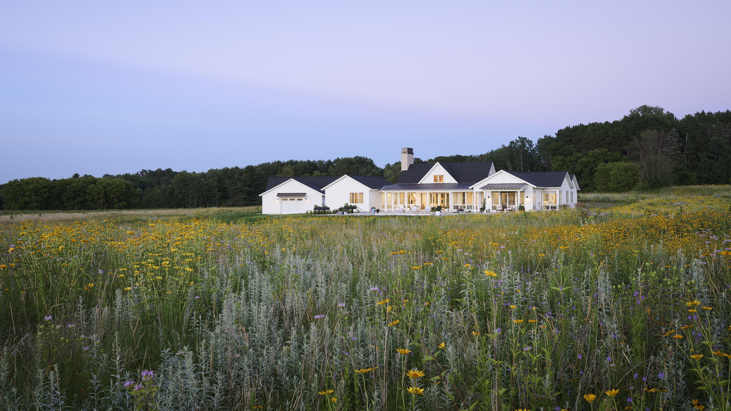 A large white house with a porch and chimney, surrounded by a field of yellow and purple wildflowers, with a dense green forest and a clear sky in the background.