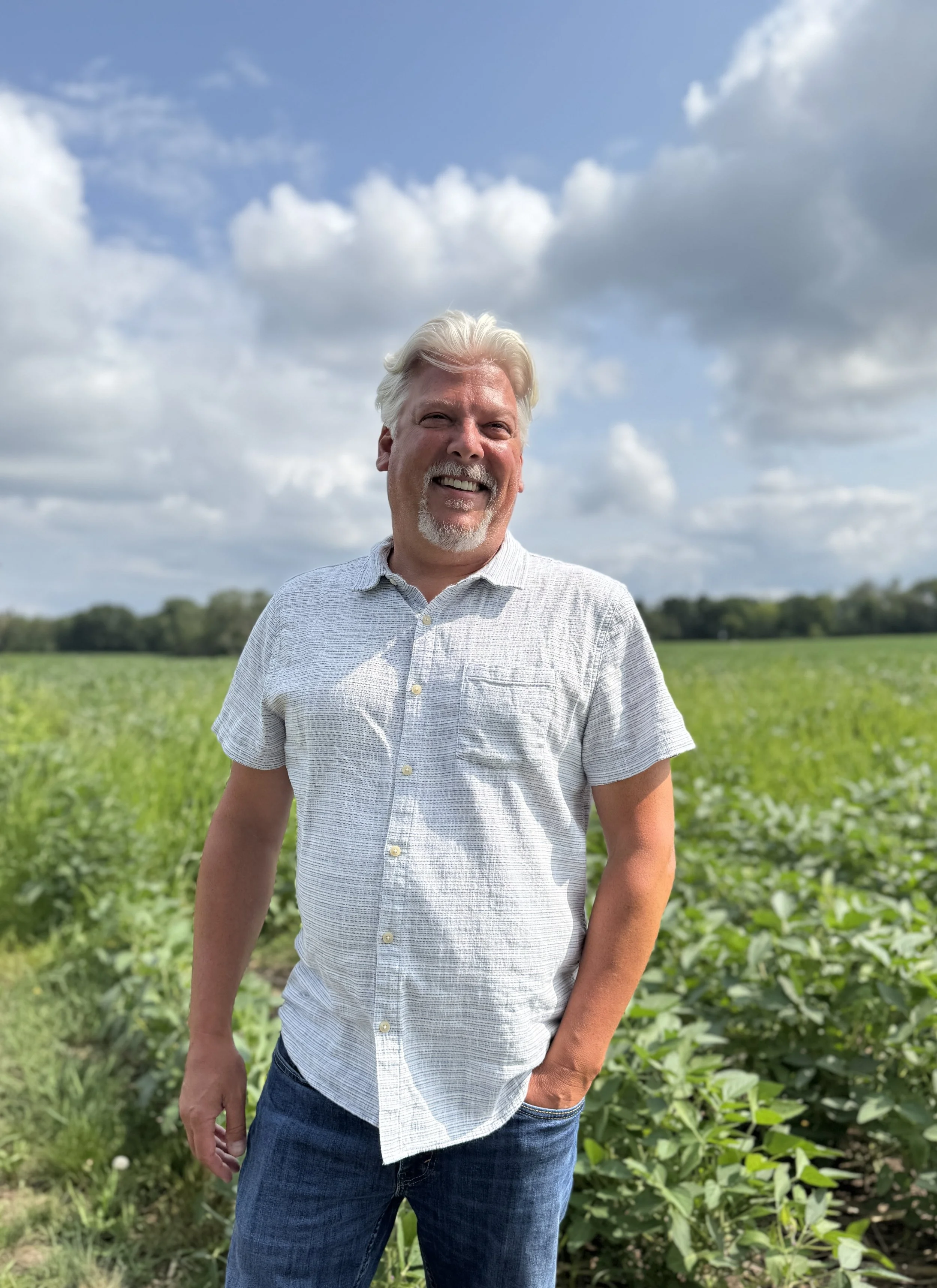 A middle-aged man with gray hair and a beard, wearing a light-colored short-sleeve button-up shirt and dark jeans, standing outdoors in a field with green plants, under a partly cloudy sky.