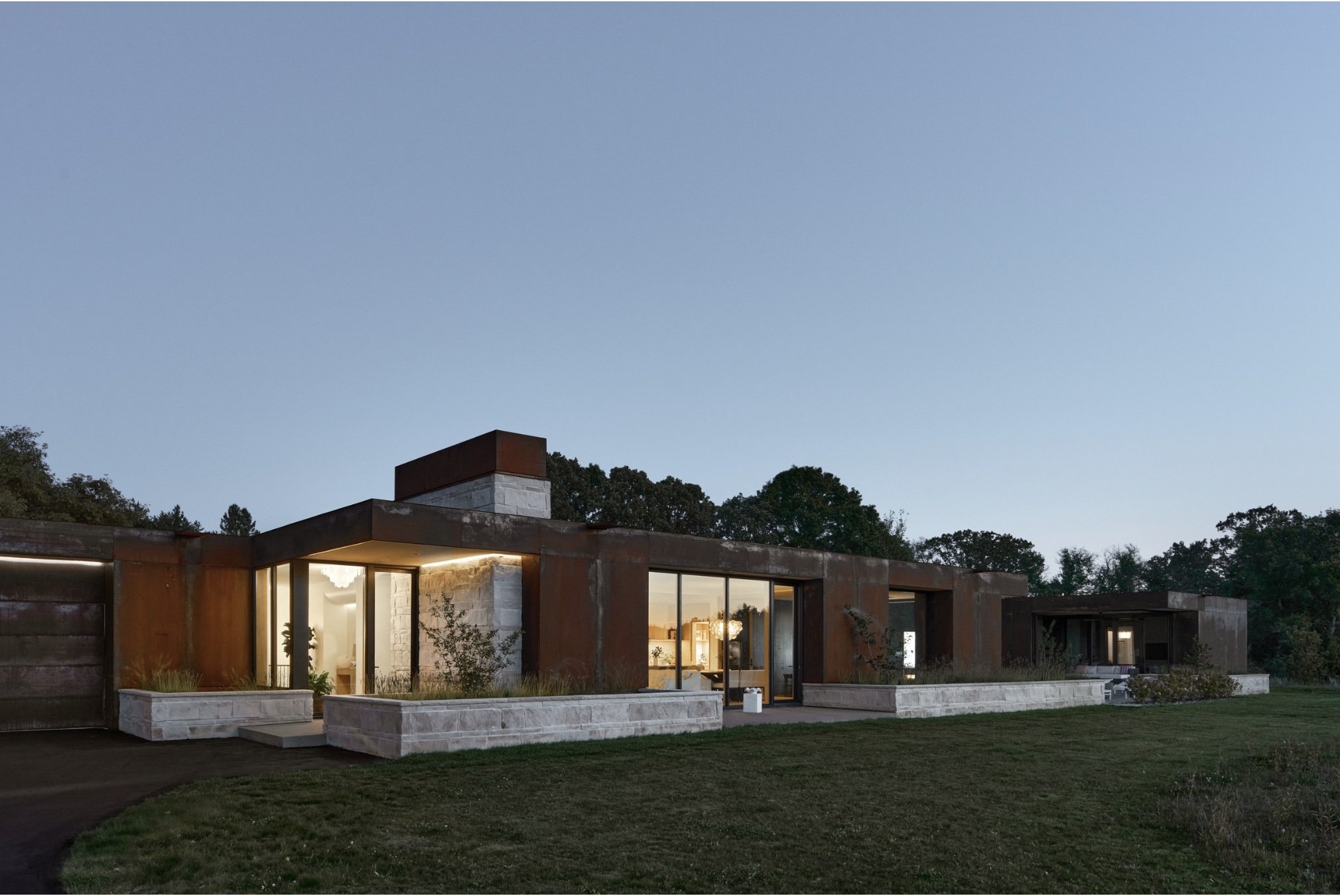 Modern house at dusk with large glass windows, stone and rust-colored exterior, surrounded by a grassy yard with trees in the background and a clear evening sky.