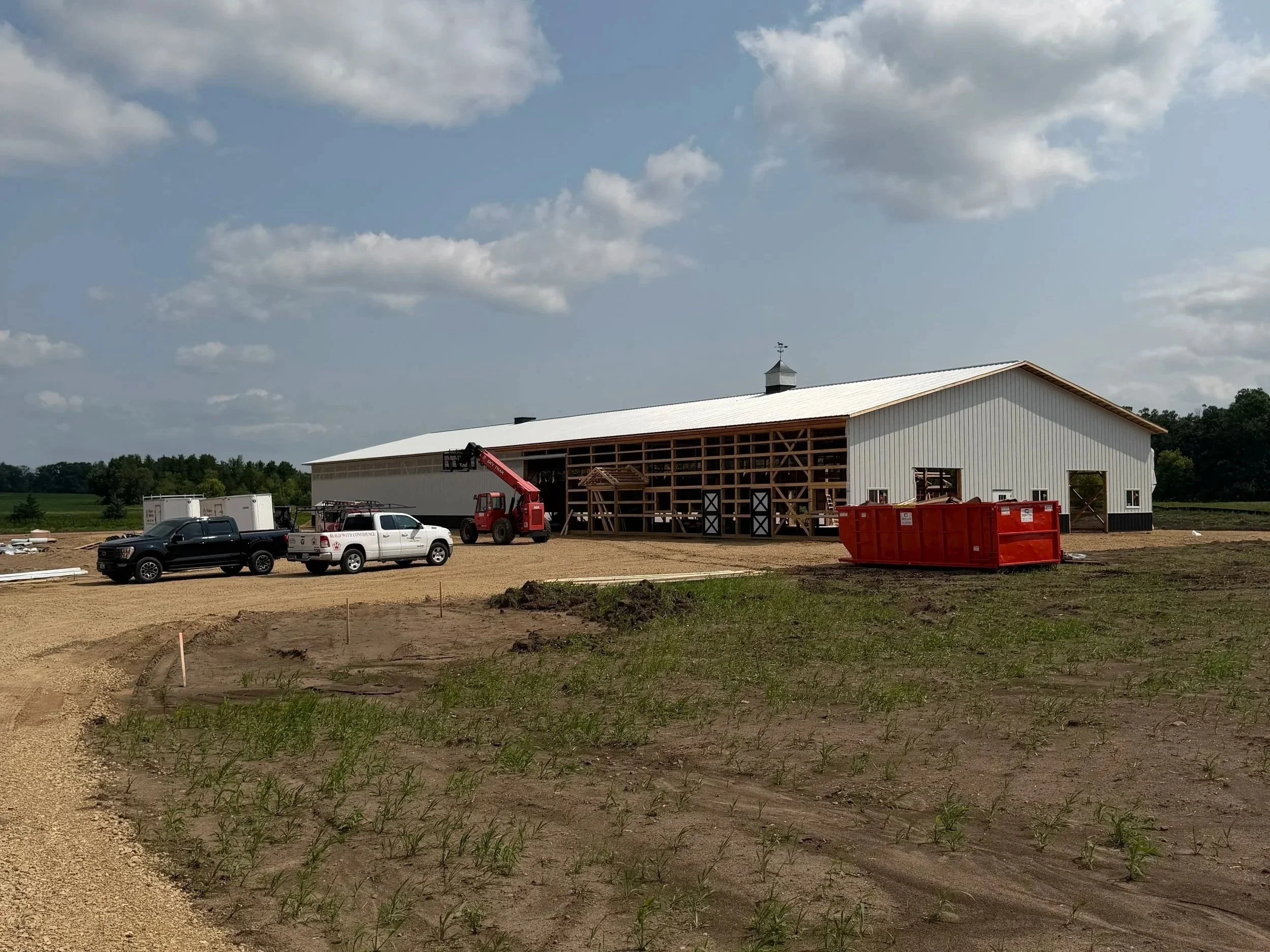 A large barn under construction with a white roof, surrounded by construction vehicles and equipment on a gravel lot with a partly cloudy sky.