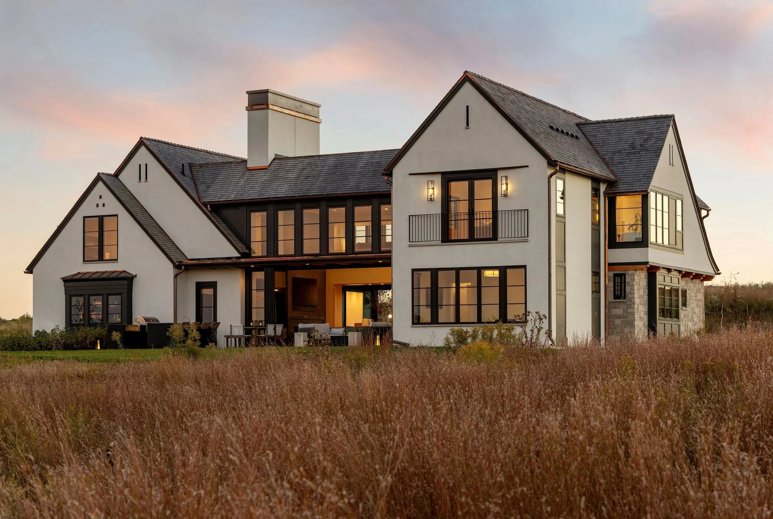 Modern two-story house with large windows and a chimney, surrounded by grassy field at sunset.