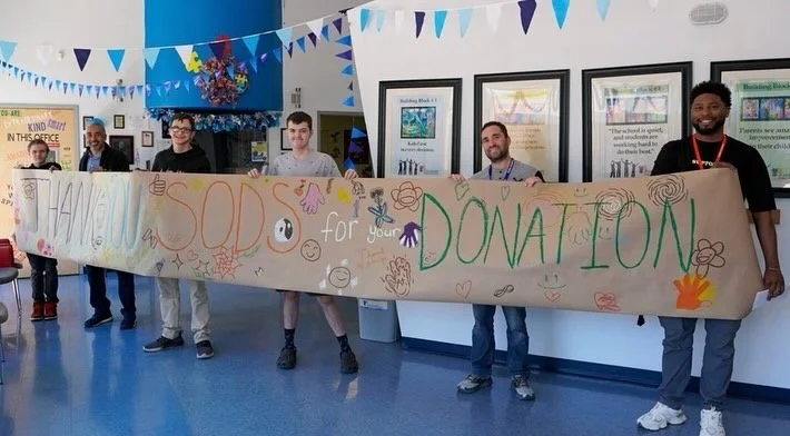 Five people holding a large handmade sign that reads 'Thankful SODA for your Donation' with colorful drawings of hearts, flowers, and smiley faces, inside a decorated indoor space with blue and white bunting.