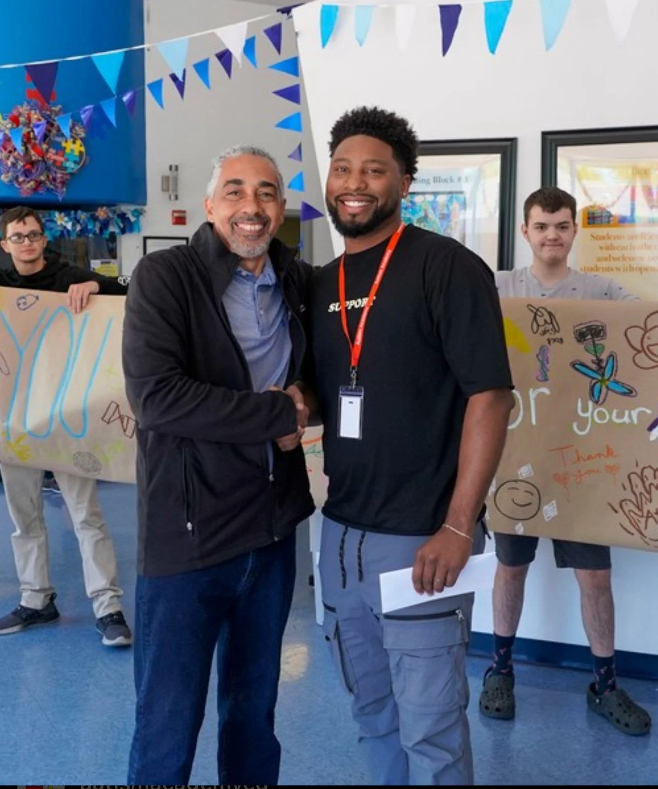 Two men shaking hands and smiling at the camera in a decorated indoor space, with banners and children holding a large poster in the background.