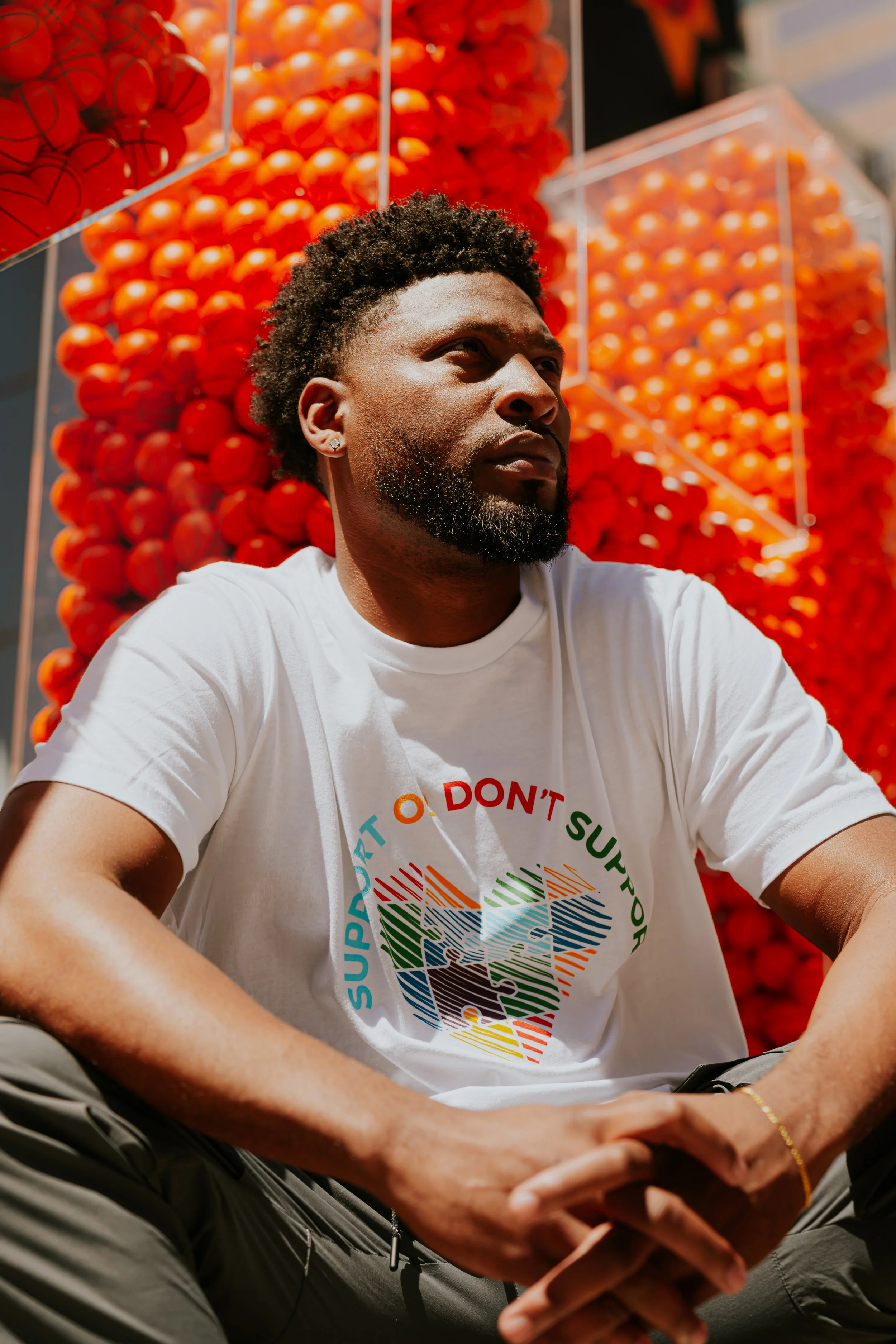A man sitting outdoors in front of a display of basketballs, wearing a white T-shirt supporting Autism Awareness Month and the text 'Support or Don't Support.