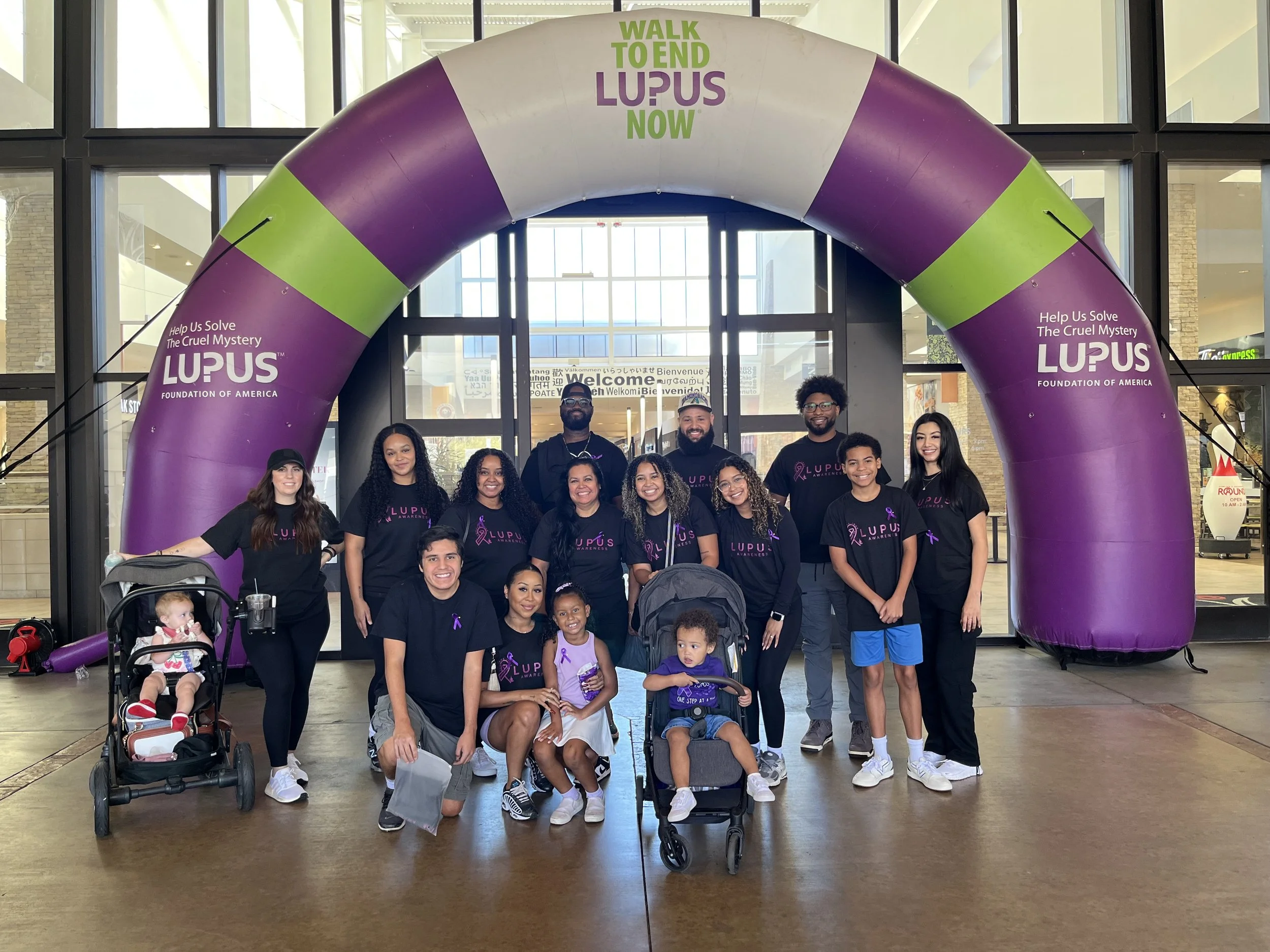 Group of people posing inside a building under a large purple and green arch with feelings of community and awareness for Lupus research. Many individuals are wearing Lupus awareness shirts with purple ribbons.