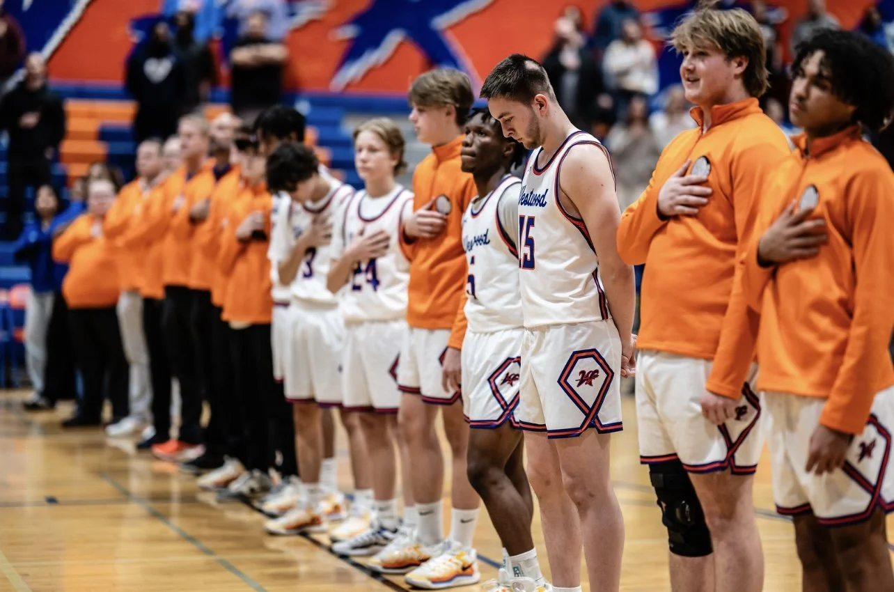 A group of basketball players and team staff standing in a line on a basketball court, with some players placing their hands on their hearts.