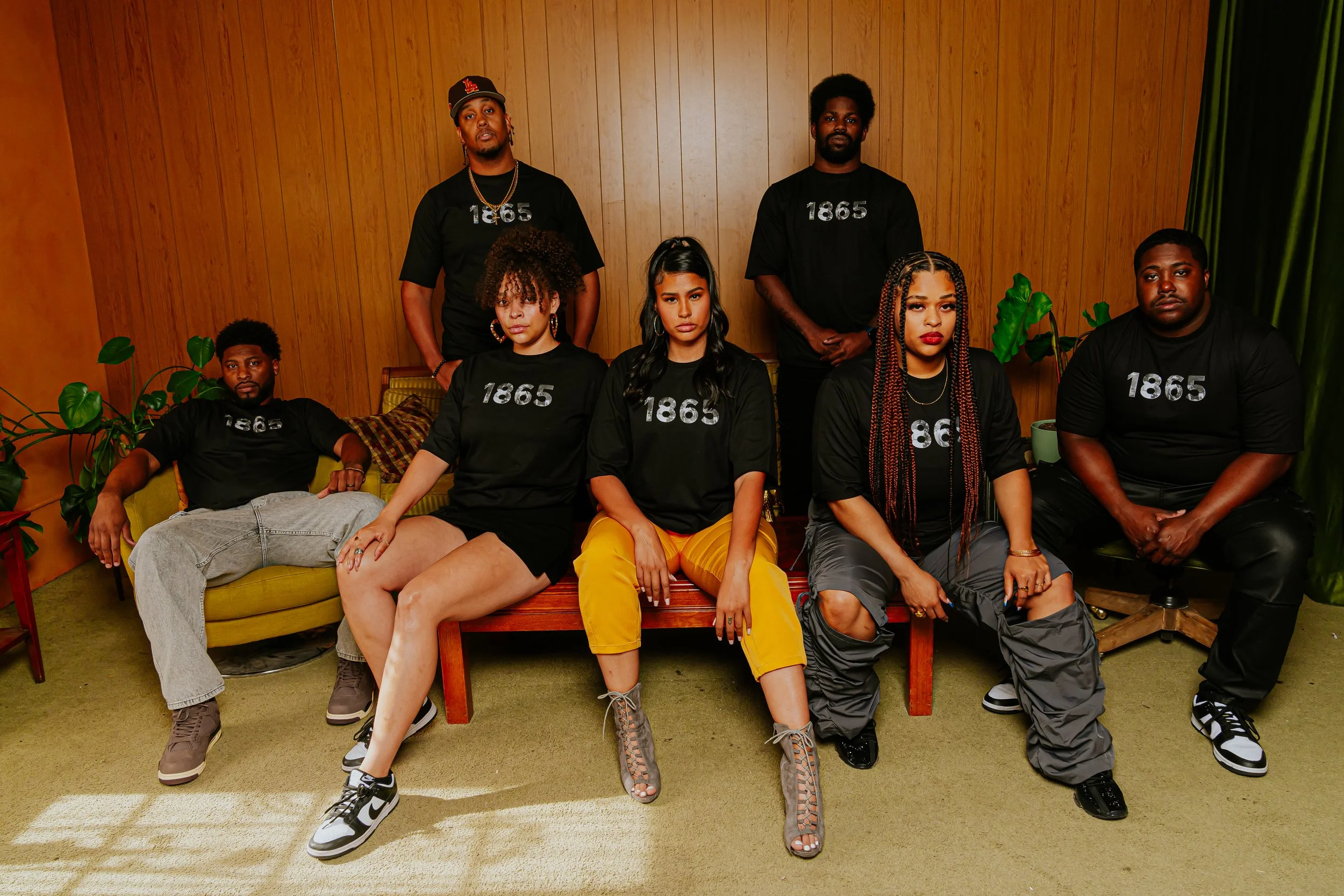 A group of seven diverse young adults wearing matching black shirts with the year '1865' printed on them, posed in a vintage-style room with wood-paneled walls, green plants, and a wooden bench.