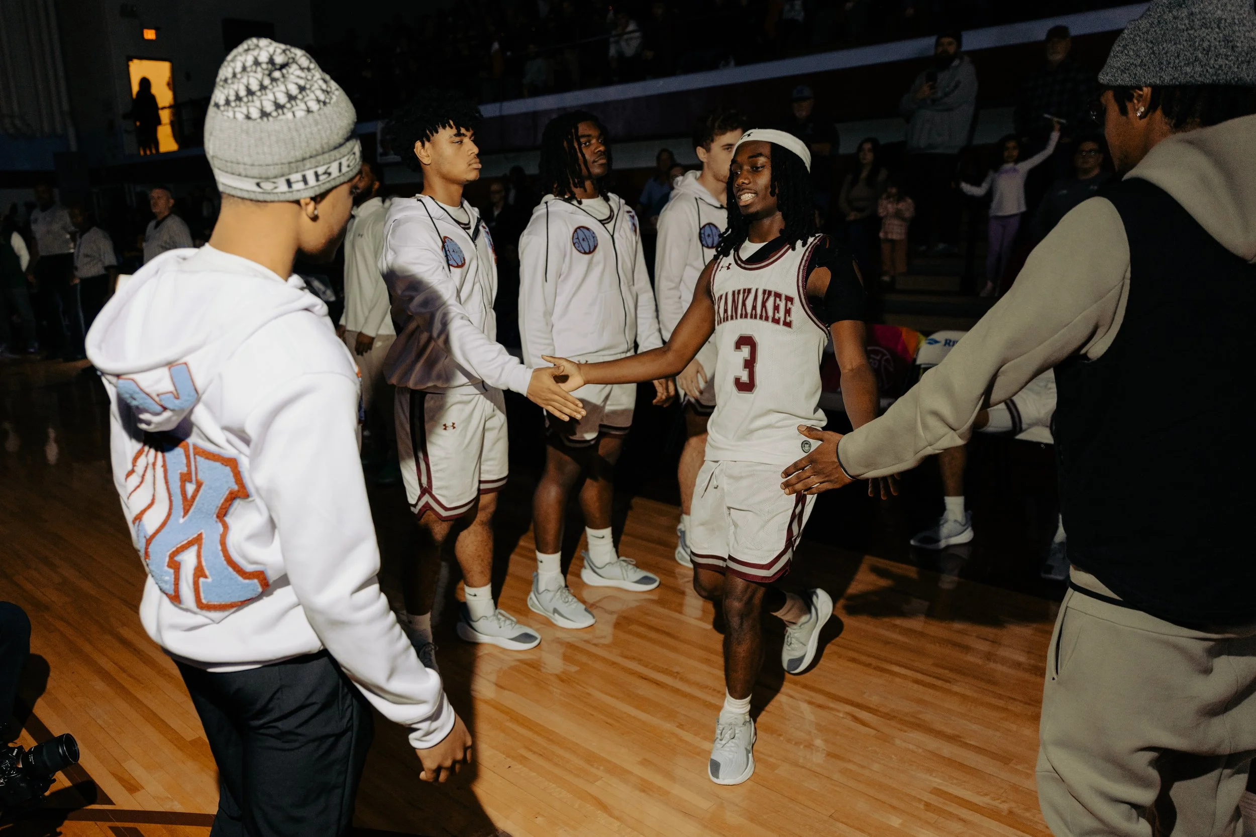 Youth basketball players on a court, one in a Kaneakee jersey number 3, shaking hands before or after a game with spectators in the background.