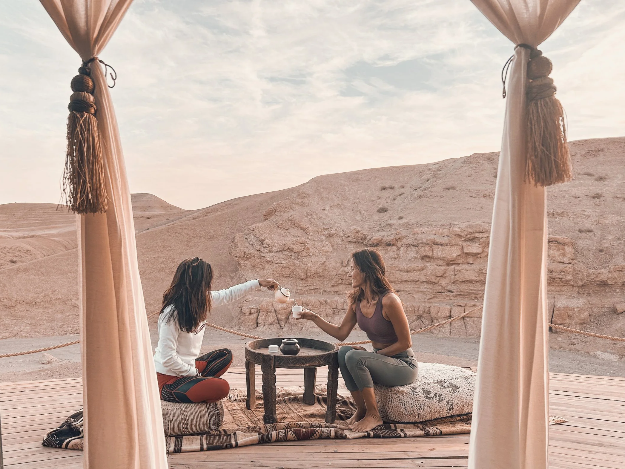 Dos mujeres sentadas en un alféizar de madera que ofrecen té en una escena al aire libre en un desierto con formaciones rocosas y cielo nublado, rodeadas por cortinas beige