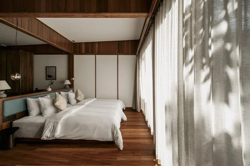 Habitación con cama grande, cortinas blancas, paredes de madera y decoraciones sencillas.