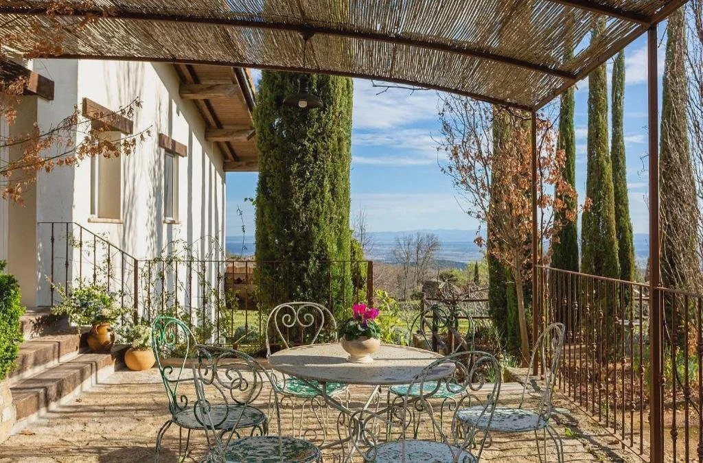 Vista de un patio con mesa y sillas de hierro, rodeado de árboles y plantas, con un toldo de paja en la parte superior y un paisaje de campo y montañas al fondo.