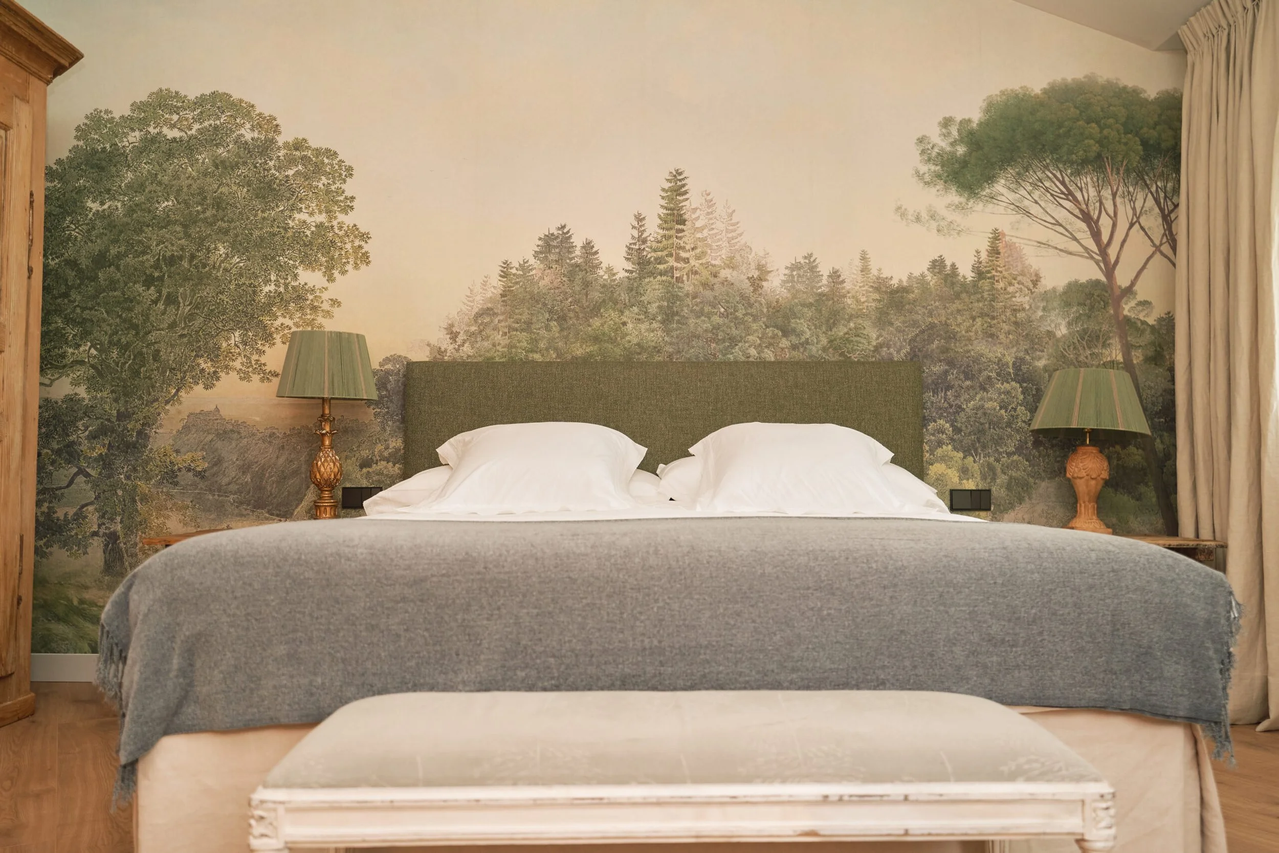 Habitación con mural de árboles en la pared, dos lámparas de noche de base de cerámica, cama con cabecera verde, ropa blanca y una colcha gris, cortinas claras a la derecha.