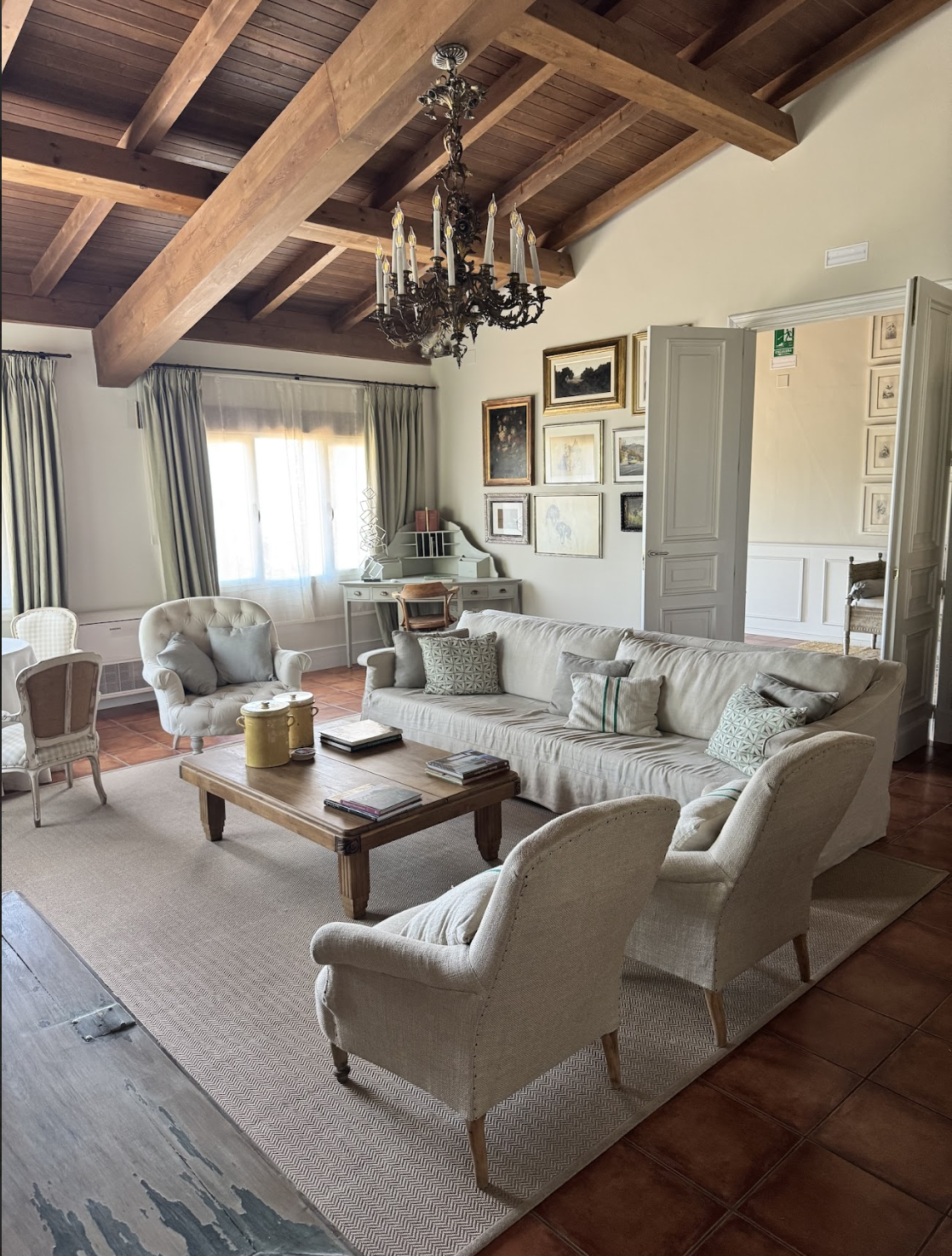 Sala de estar con techo de madera, lámpara de araña, paredes decoradas con cuadros y sofás blancos con cojines, mesa de centro de madera y ventanas con cortinas beige.