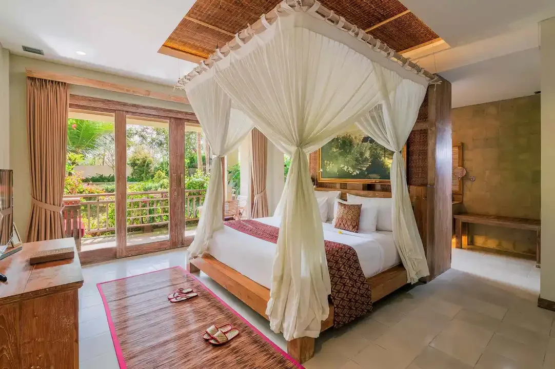 Habitación con cama de dosel, vista a un balcón con vegetación, cortinas beige, y decoración en tonos madera y beige.